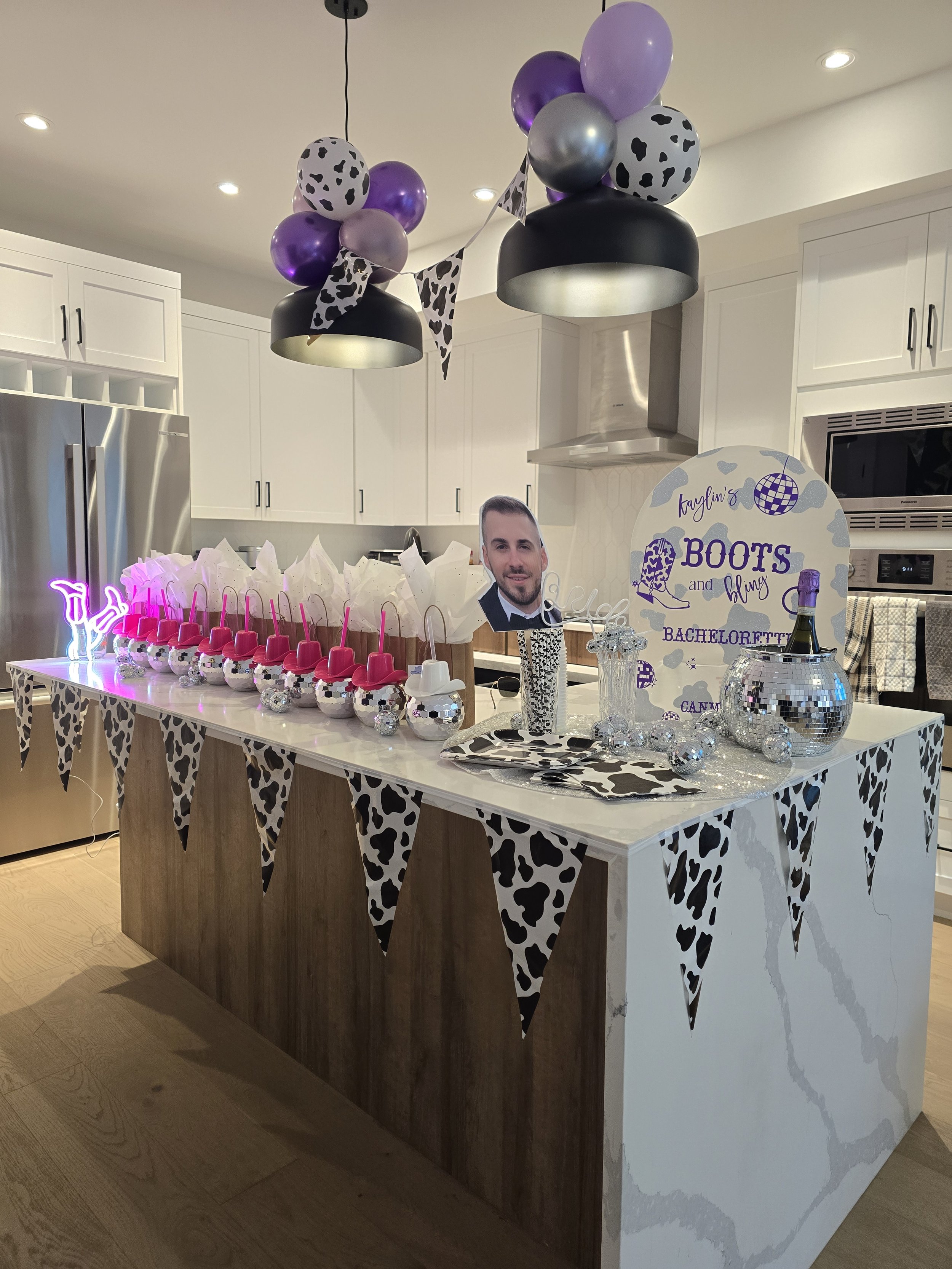 Party setup with black, purple, and cow print balloons hanging from the ceiling. A decorated table features pink cups with straws, with cow print and black and white themed decorations. A sign reads 'Kaylin's Boots and Bling Bachelor Party.' There is a large silver disco ball, a picture of a man, and a champagne bottle on the table in a silver holder.