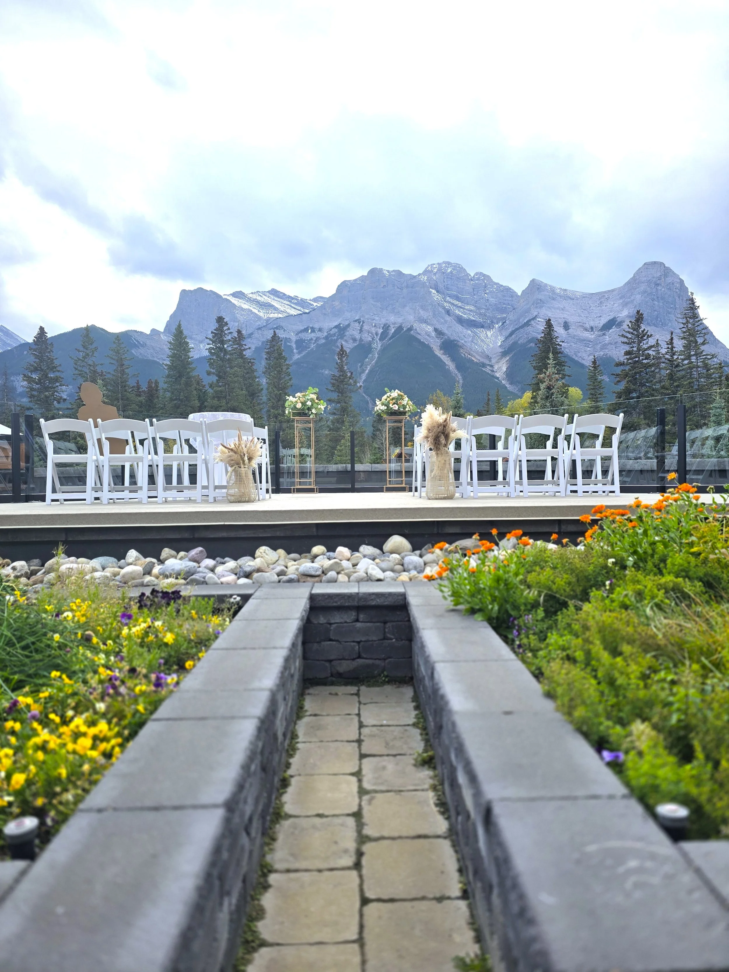 A flower bed with colorful flowers on either side of a walkway. In the background, outdoor chairs and tables, flower arrangements, and a mountain range with snow-capped peaks and pine trees under a cloudy sky.
