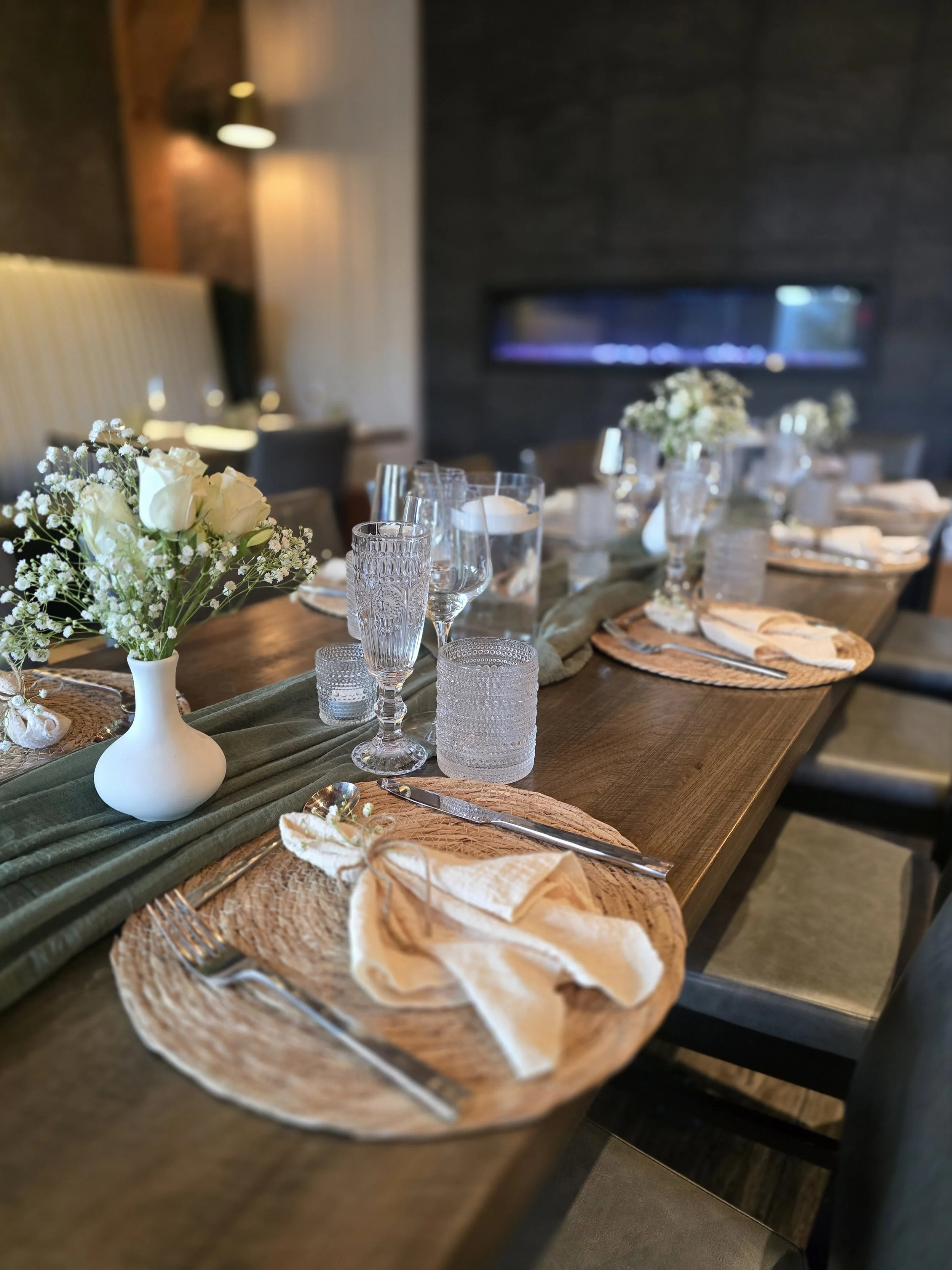 A dining table set with floral centerpieces, glassware, silverware, woven placemats, and napkins, in a room with dark walls, an electric fireplace, and warm lighting.