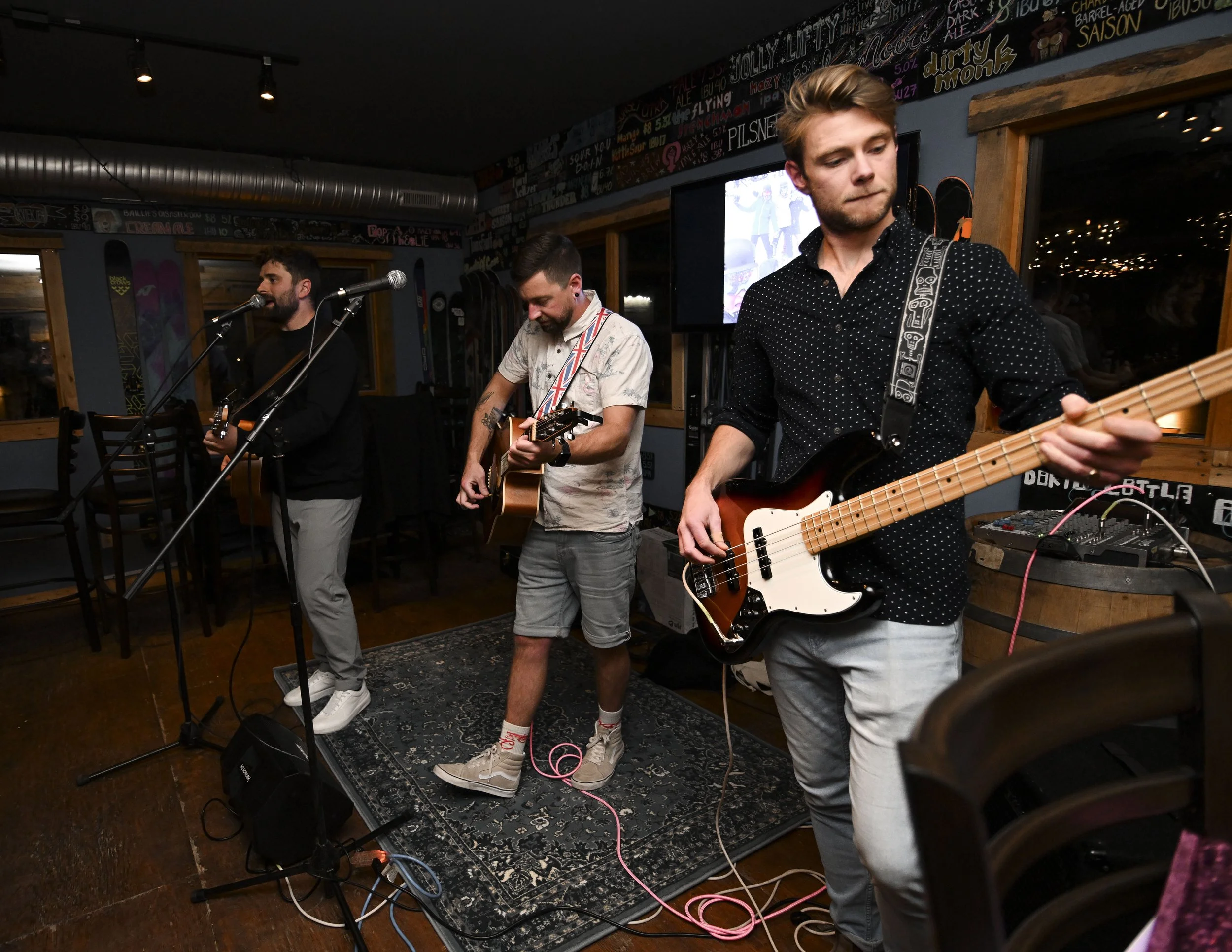 Three musicians performing at a small indoor venue with colorful artwork on the walls, featuring a vocalist with a microphone, a guitarist, and a bassist. The scene includes musical equipment and string lights reflected in the window.