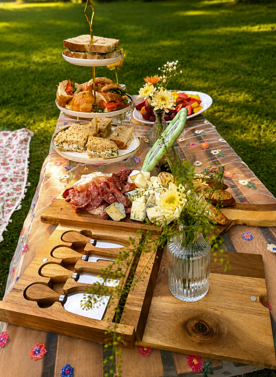 Outdoor picnic table with cheese, charcuterie, bread, fruit, and flowers on a floral tablecloth.