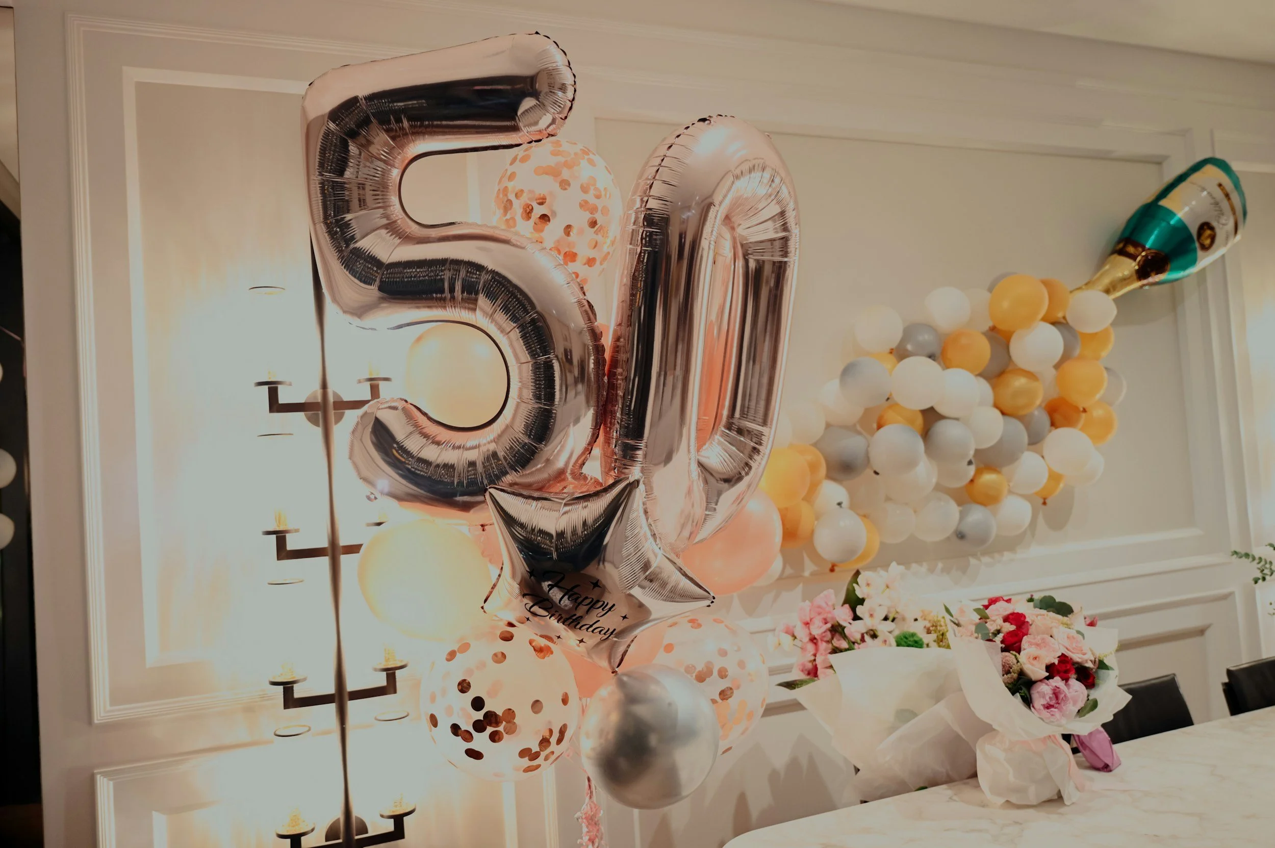 Decorative celebration setup with large metallic balloons, a bouquet of pink and dark red flowers, and a wall decorated with balloons in white, yellow, and gray.