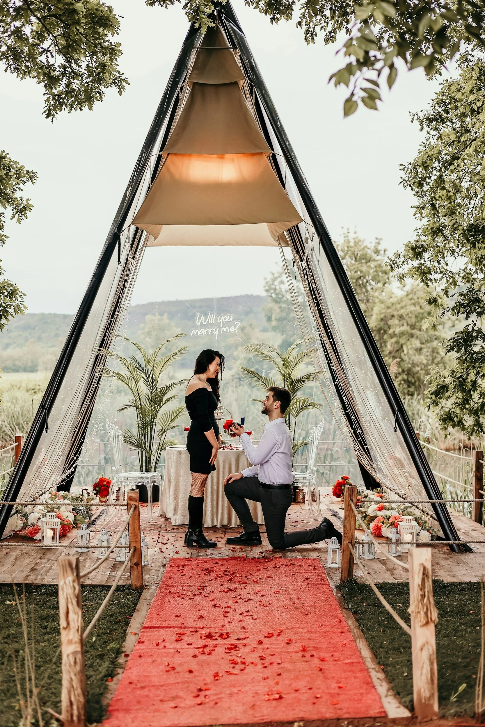 A man proposing marriage to a woman on a red carpet aisle decorated with flower petals, under a tent structure with trees in the background