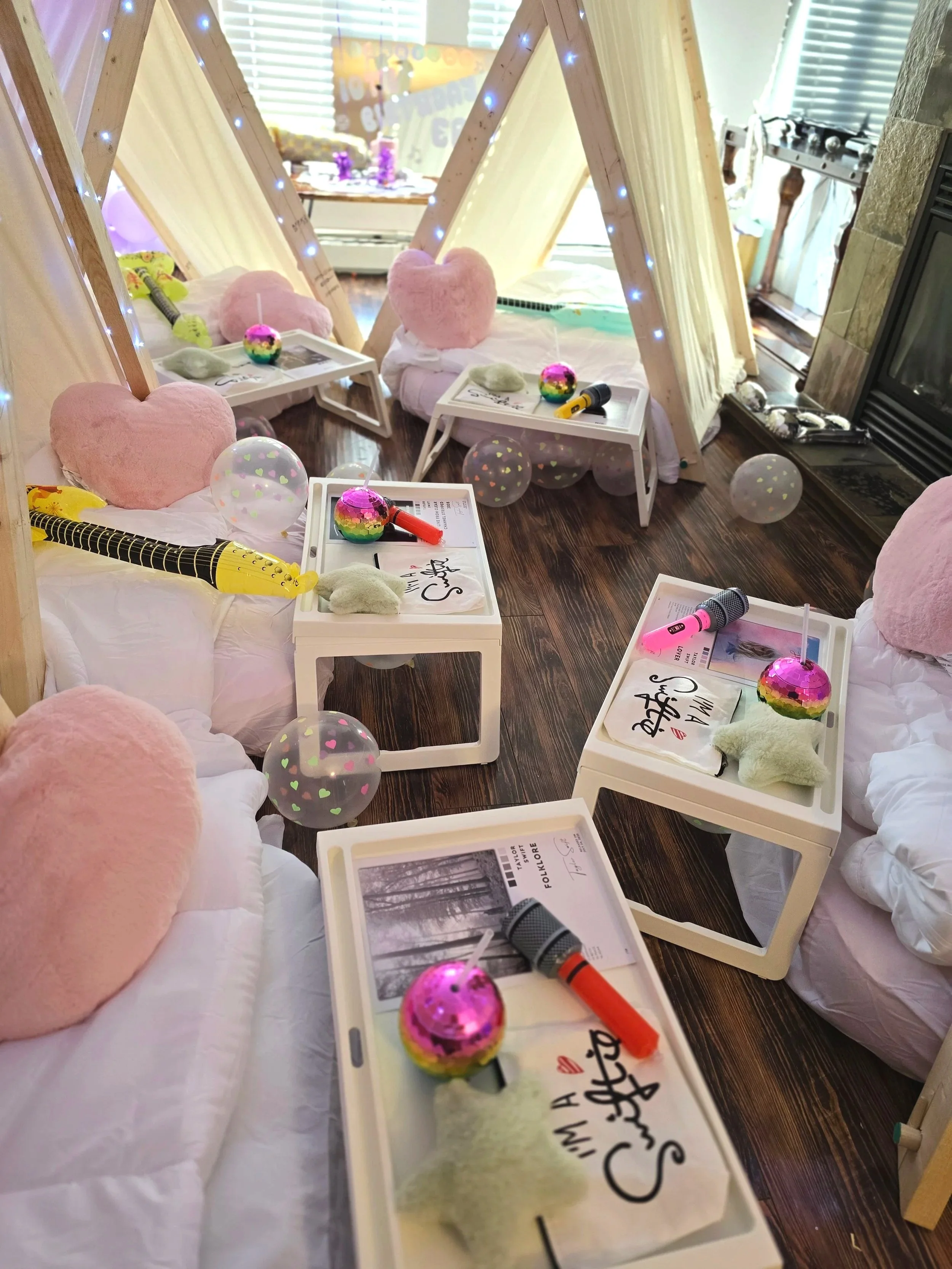 Children's indoor play tent with pink heart pillows, stuffed animals, small tables with toys, books, and flashlights, some balloons, and fairy lights, in a cozy room with a fireplace.