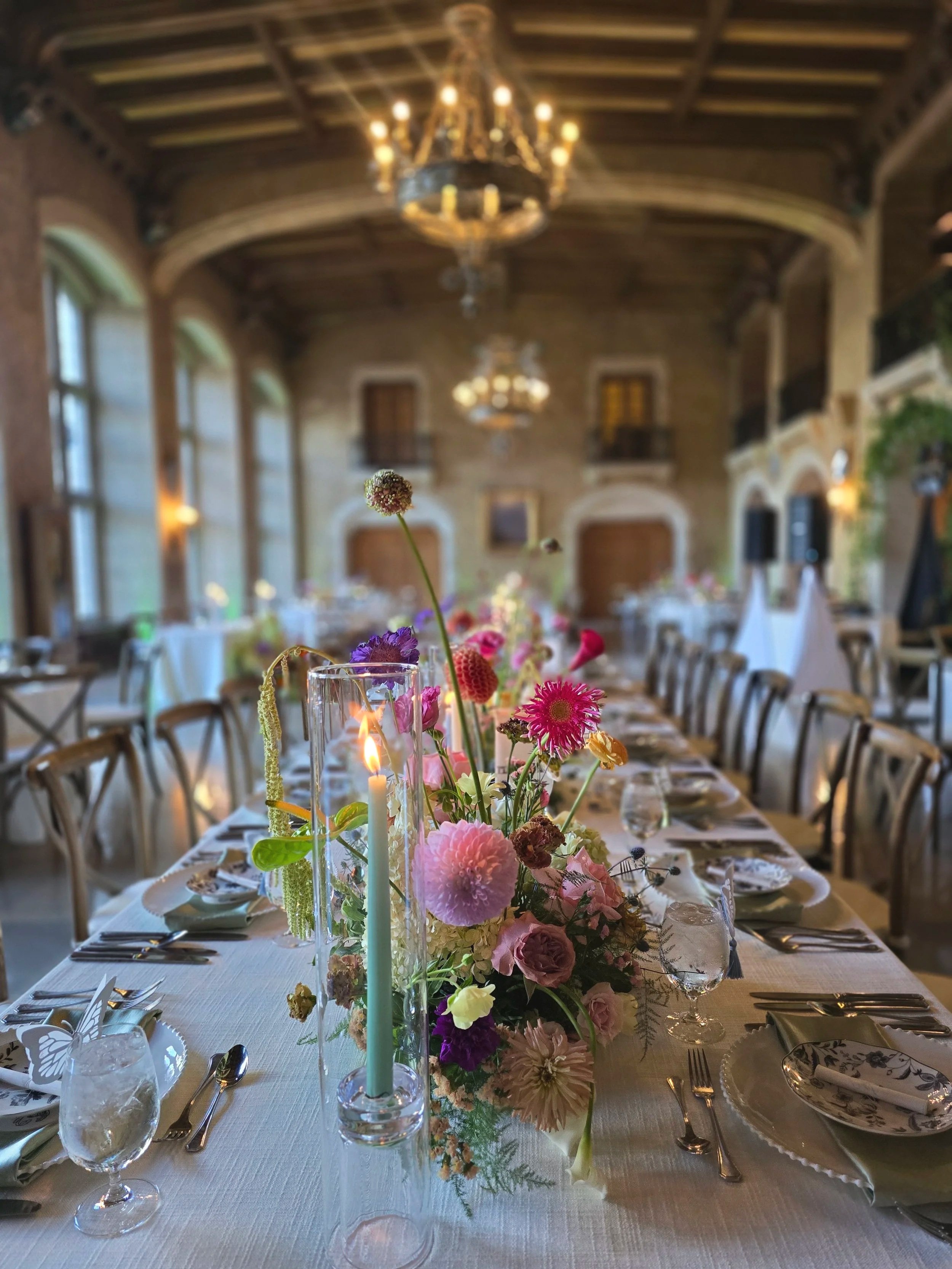 Elegant banquet table with a floral centerpiece, candles, and place settings in a spacious, well-lit room with high ceilings and chandeliers.