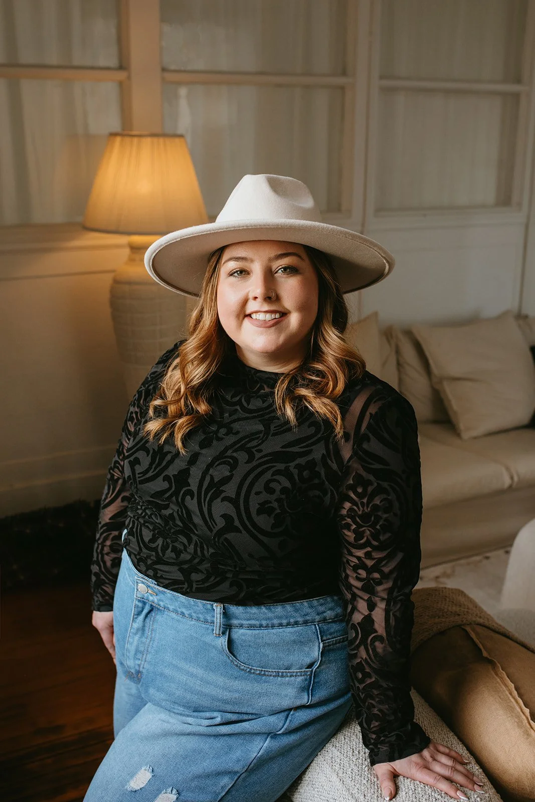 A woman with light skin and shoulder-length wavy hair wearing a wide-brimmed cream hat, a black patterned long-sleeve top, and light blue jeans, sitting on a sofa in a cozy, softly lit room with a lamp and pillows in the background.