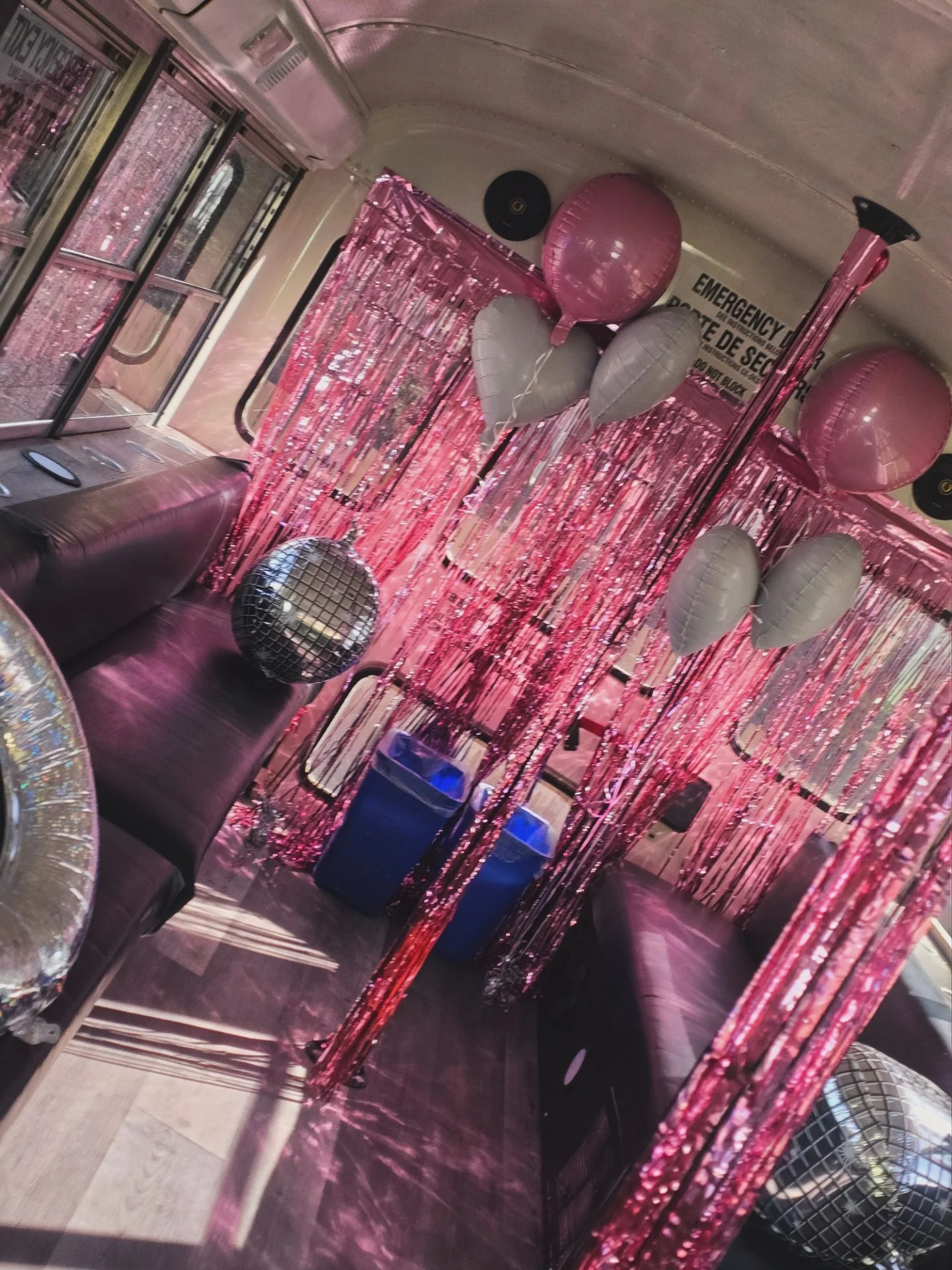 Interior of a vehicle decorated with pink tinsel streamers, heart-shaped balloons in pink and gray attached to the ceiling, a disco ball, and a sign that reads 'Emergency Exit' in English and Spanish.