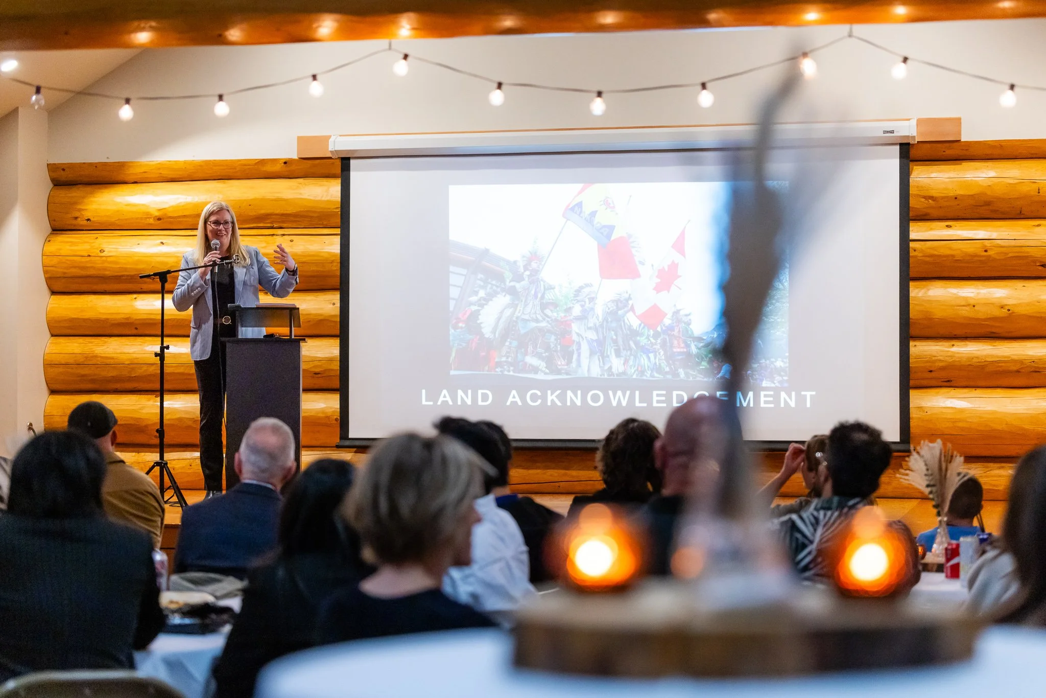 A woman giving a speech at a conference or workshop, standing at a lectern with a microphone, in front of a projection screen with the words 'LAND ACKNOWLEDGEMENT' and a graphic of Indigenous people and flags, in a room with wooden walls and string lights, with an audience seated facing her.
