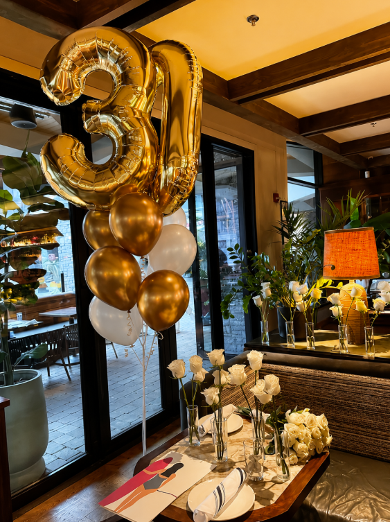 Gold and white balloons, including a large gold number 30, decorating a dining table with white roses and place settings inside a restaurant