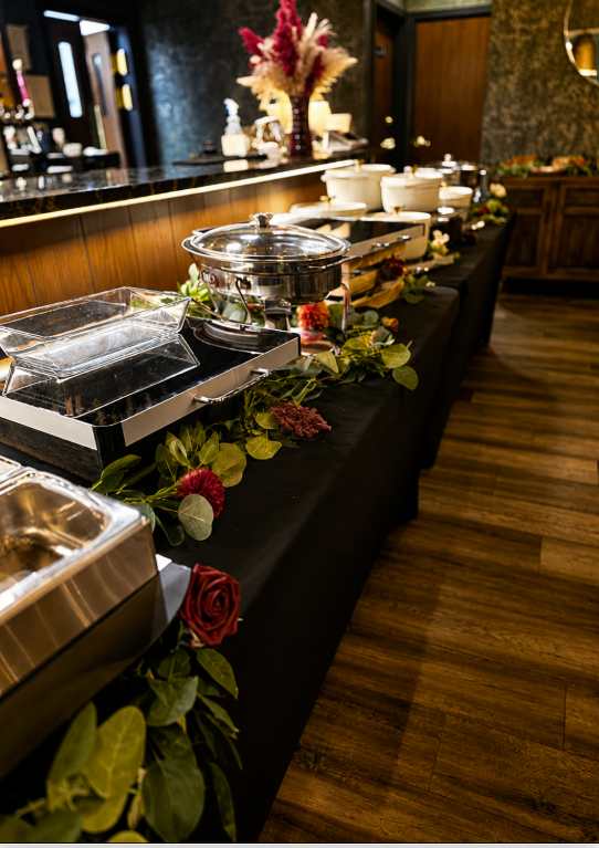 Buffet table with food containers, some covered with glass lids, decorated with greenery and roses, in a restaurant setting.