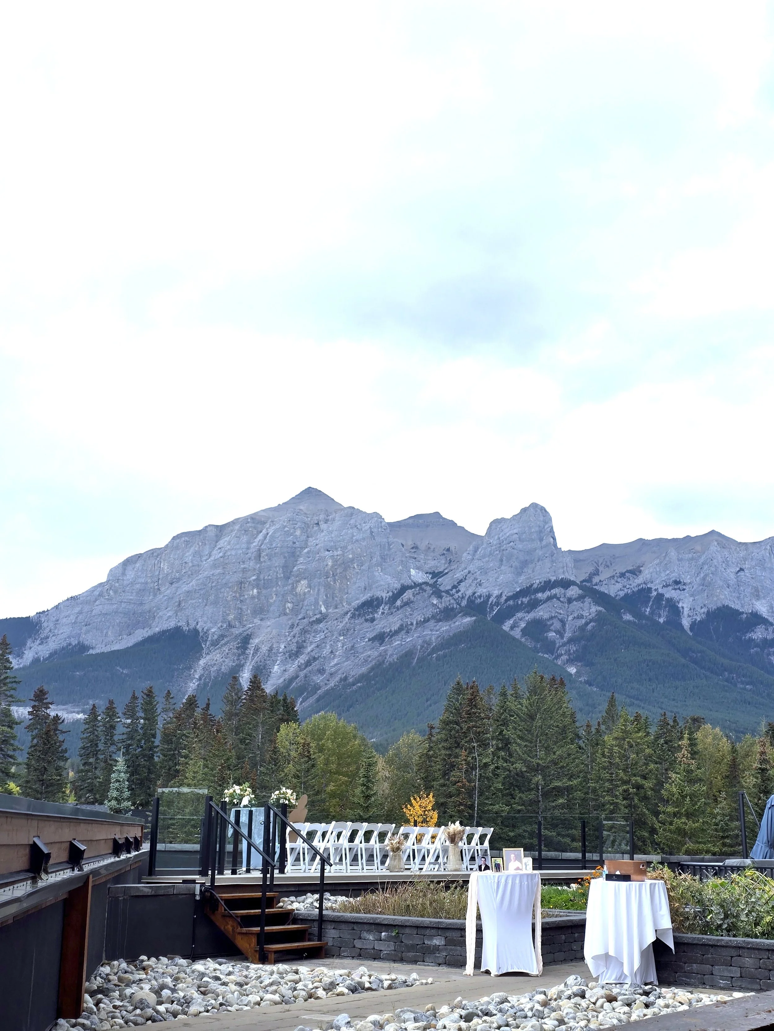 Outdoor event setup with white chairs and tables with white tablecloths, overlooking a mountain range with a cloudy sky and some trees in the background.