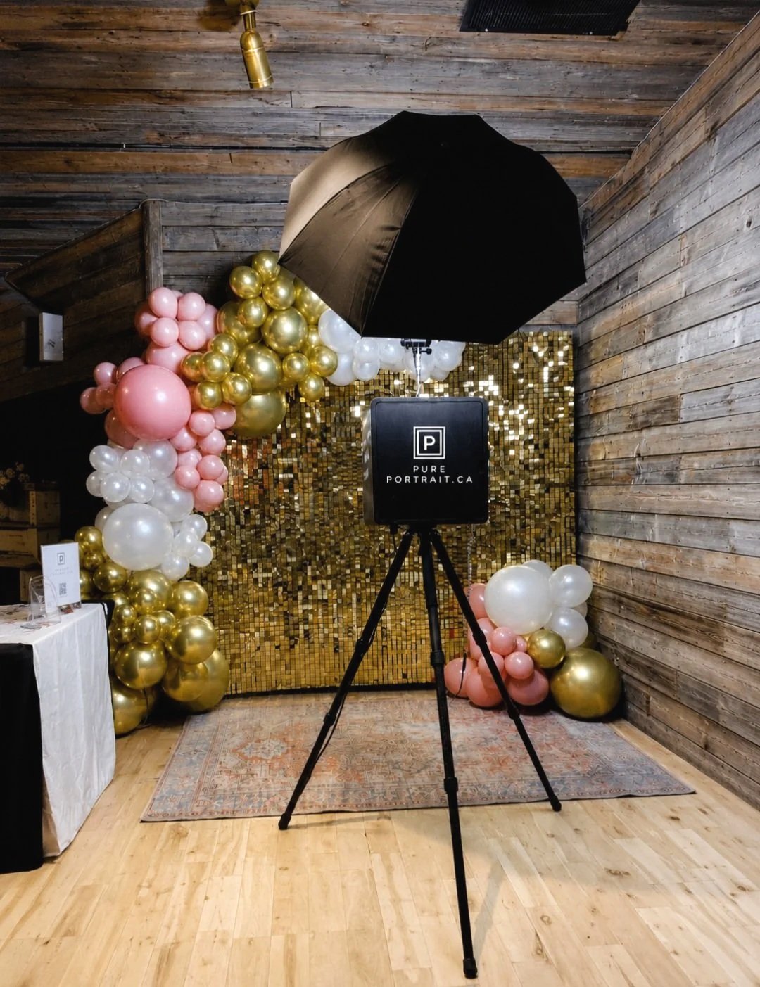 Photo of a photo booth setup with a backdrop of gold mosaic tiles, decorated with pink, white, and gold balloons, a black umbrella for lighting, and a professional camera on a tripod.