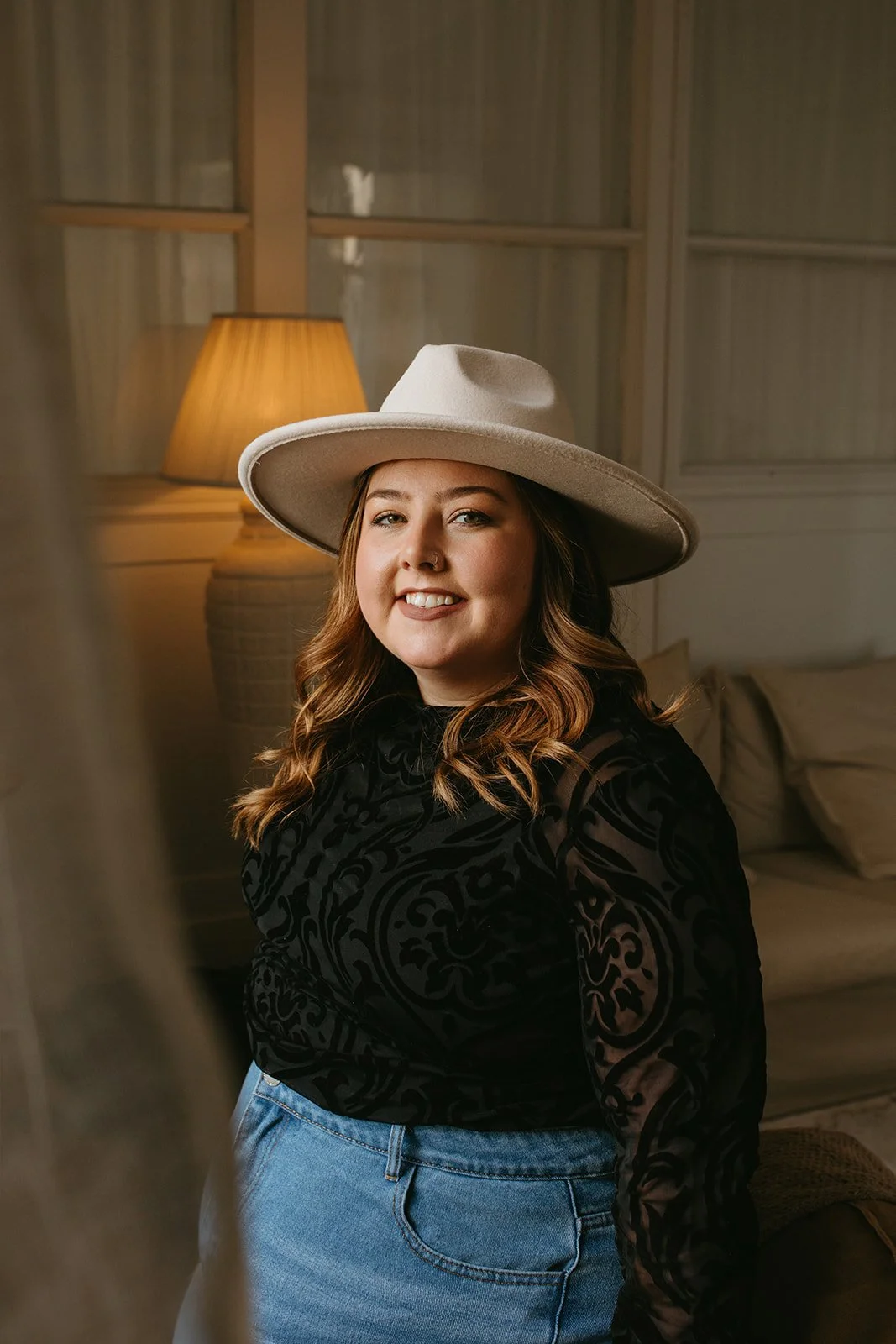A woman with wavy brown hair, wearing a beige wide-brimmed hat and a black patterned blouse, smiling indoors with a lamp and sofa in the background.