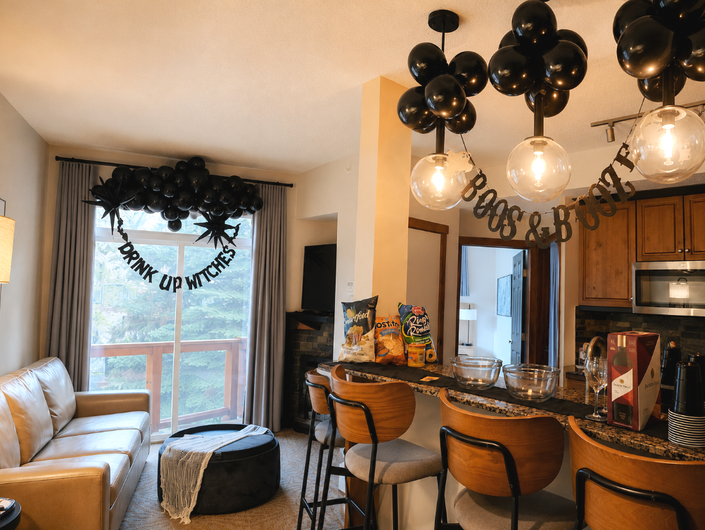 Living room with Halloween decorations including black balloons and banners that say "Drink Up Witches" and "Boo & Bost". There is a beige sofa, a black pouf, a TV, and a kitchen with barstools and snacks.