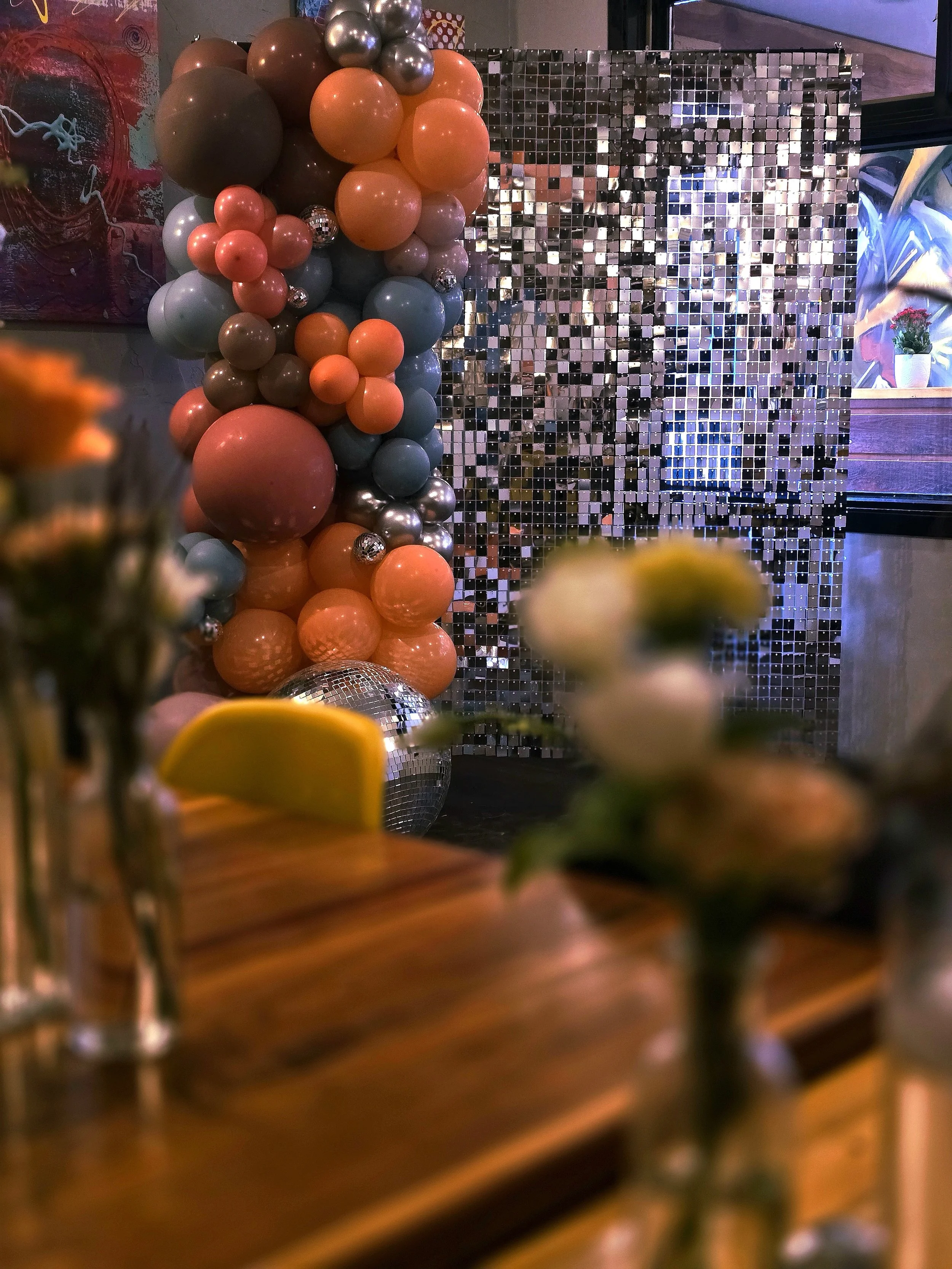 A decorative arrangement of balloons in various sizes and colors, including coral, blue, gray, and metallic silver, next to a reflective mosaic wall, with blurred objects and flowers in the foreground.