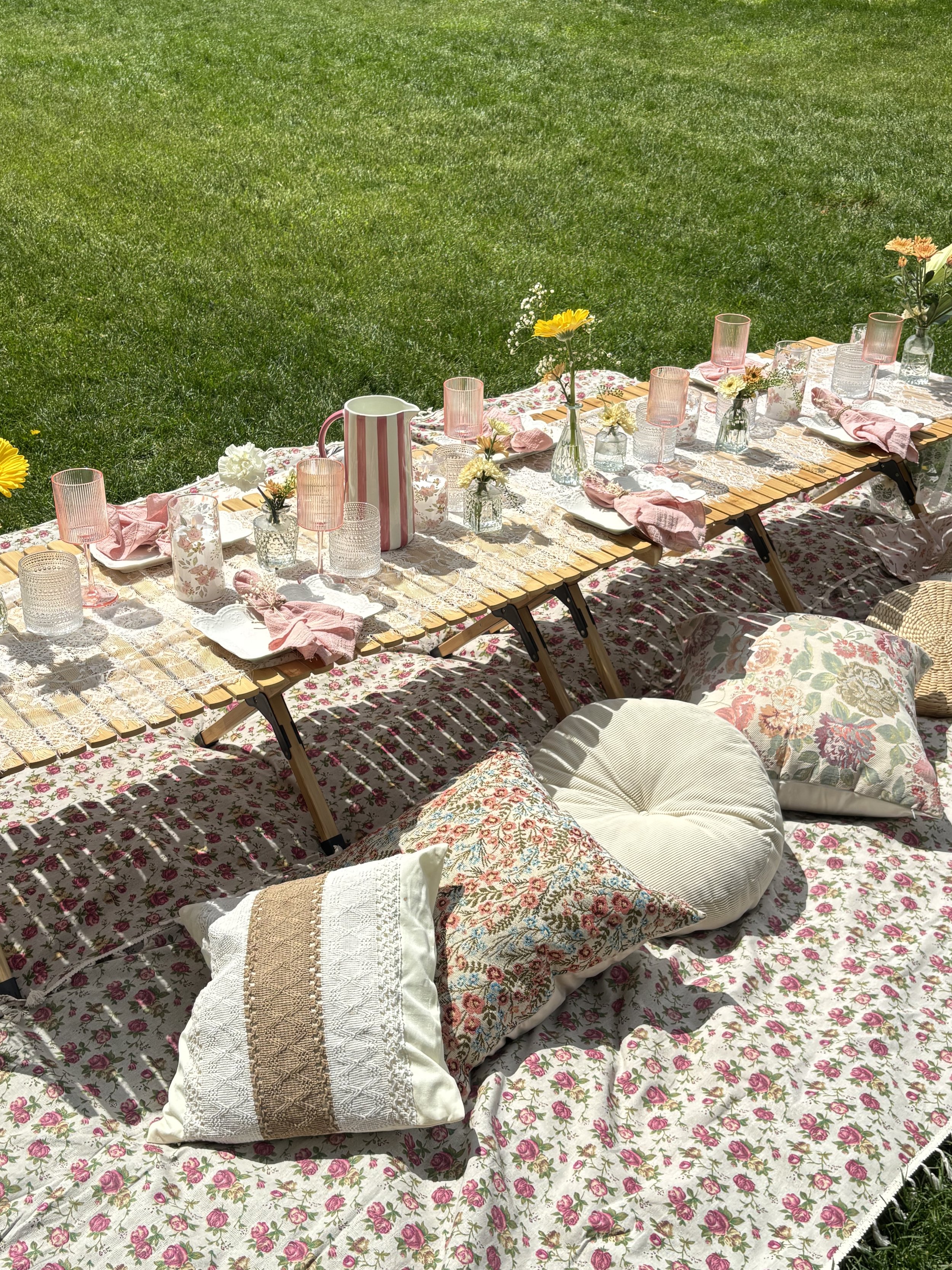 An outdoor table set with floral tablecloth, pink glasses, and vases of flowers on a grassy lawn.