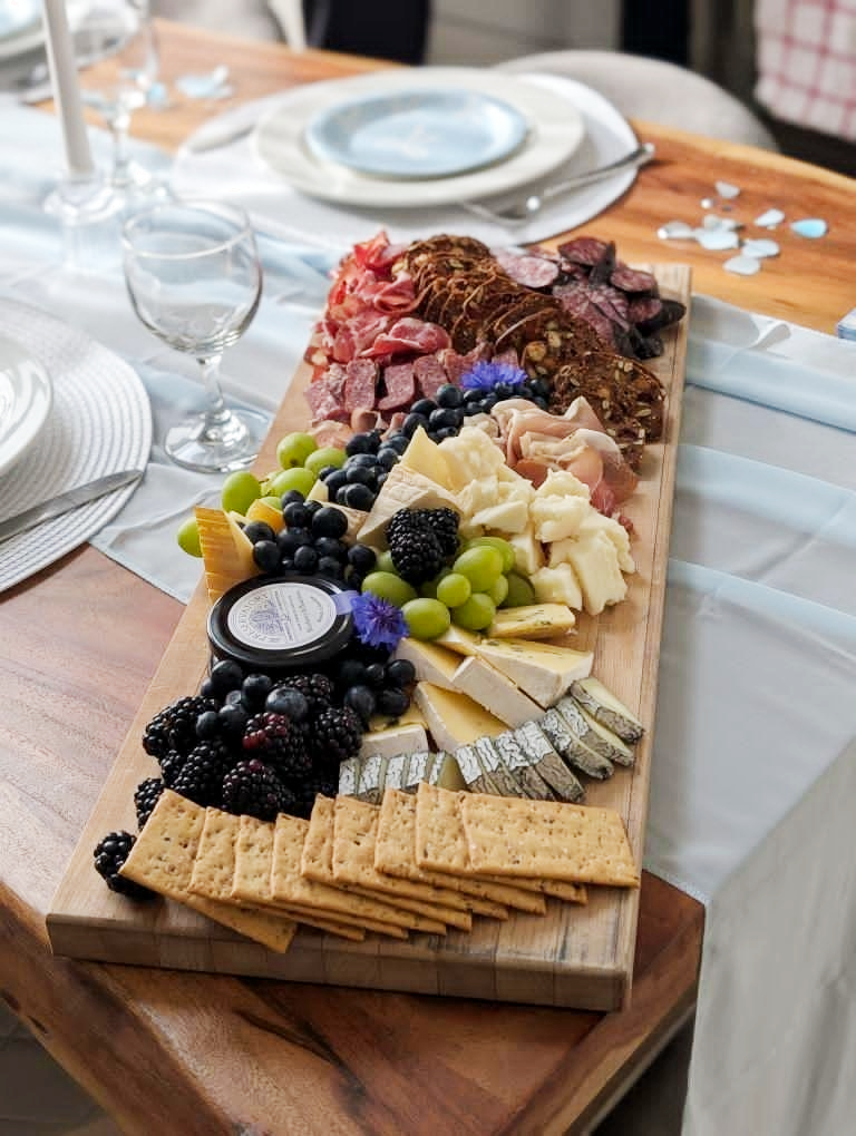 Charcuterie board with assorted cheeses, meats, grapes, berries, crackers, and a small jar of honey on a wooden table with plates and glasses nearby.
