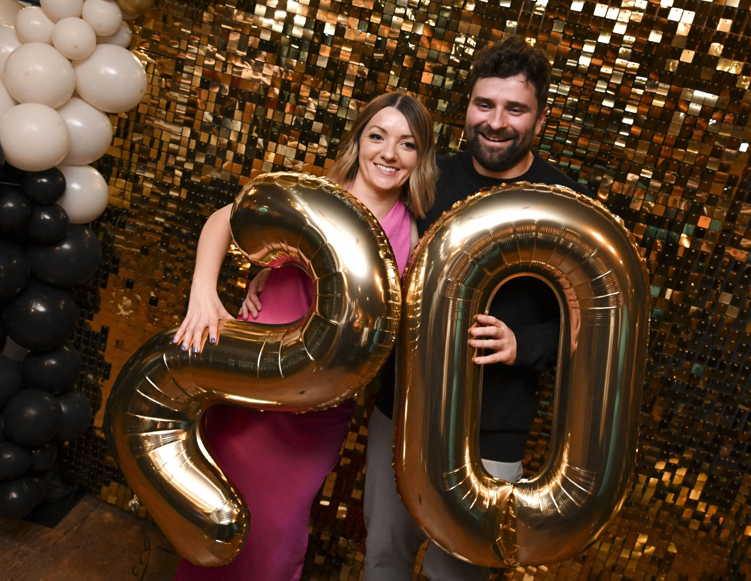 A woman in a pink dress and a man in a black shirt smiling and holding large gold balloon numbers 2 and 0 at a party with gold and black decorations and a mosaic gold and black background.