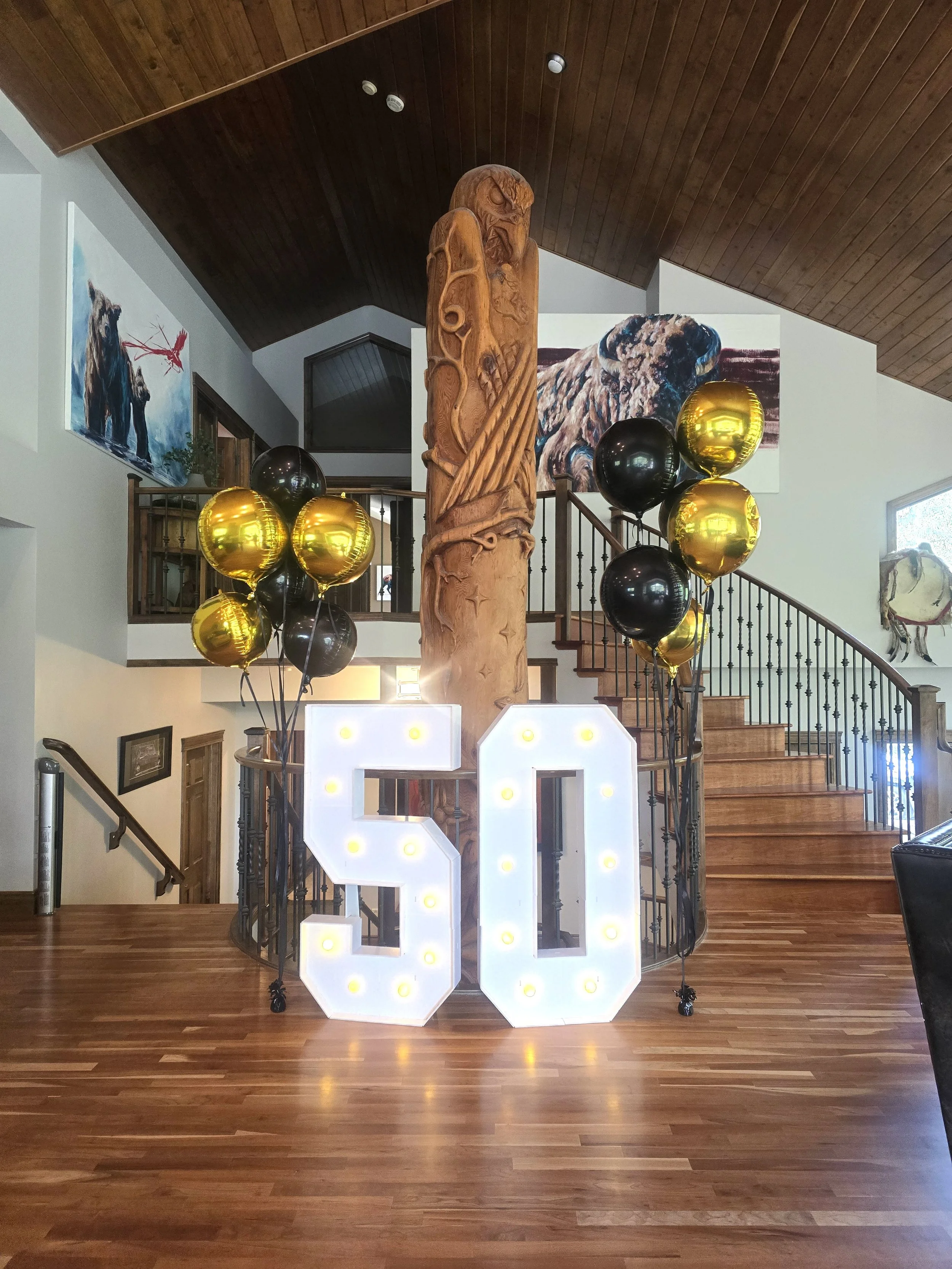 Indoor celebration setup with illuminated white letters spelling '50', gold and black balloons, a carved wooden totem pole, two large wildlife paintings on the wall, and wooden stairs.
