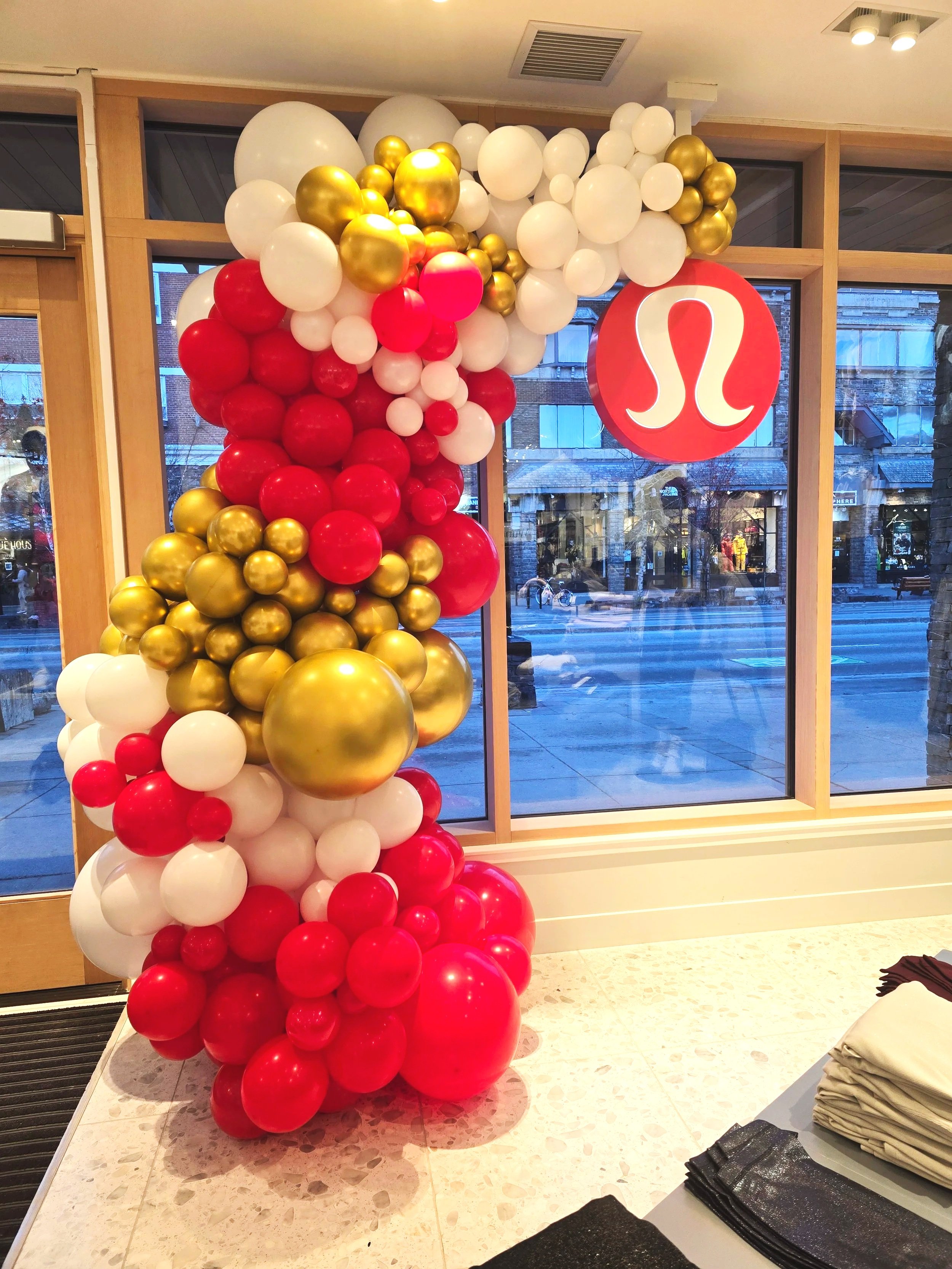 Decorative balloon arrangement with red, white, and gold balloons near a window, featuring a red Lululemon sign.