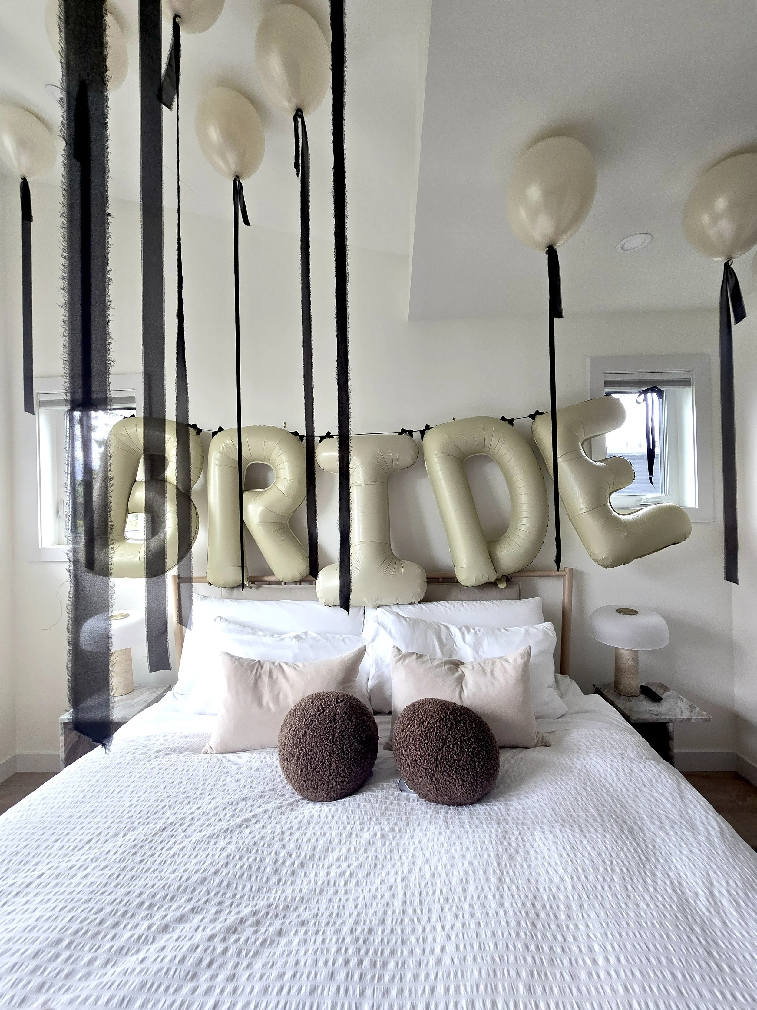 A bedroom with a bed covered in a white textured bedspread and three brown round pillows. Above the bed, large beige balloon-like letters spell 'LOVE'. Black ribbons and white balloons hang from the ceiling. There are two small windows with blinds, and a small nightstand with a lamp on it.