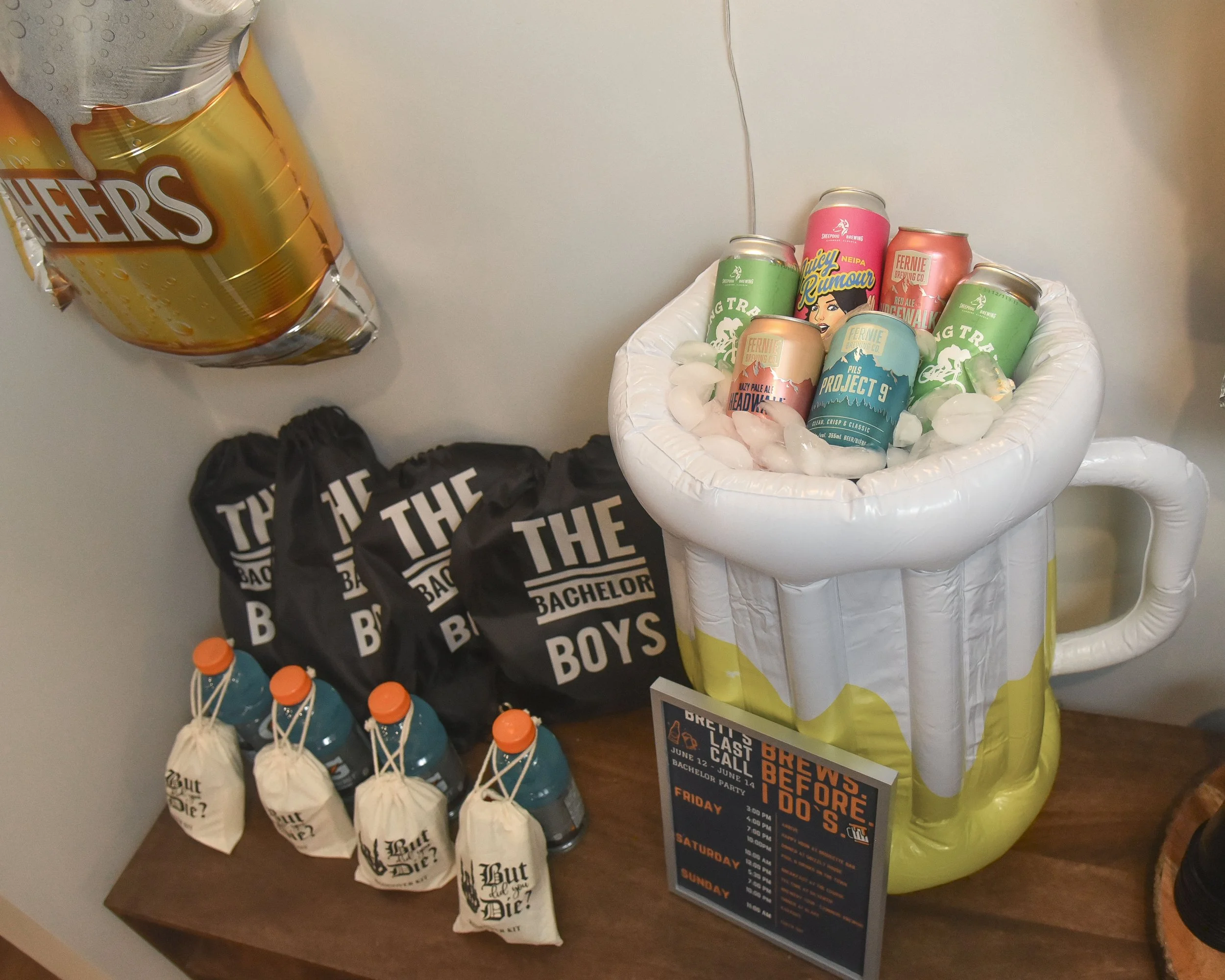 A display of canned beers in a white ice bucket, with colorful beer cans featuring various designs, a large balloon shaped like a beer mug, black tote bags with white lettering, and bottles of orange liquid, likely soda or juice.
