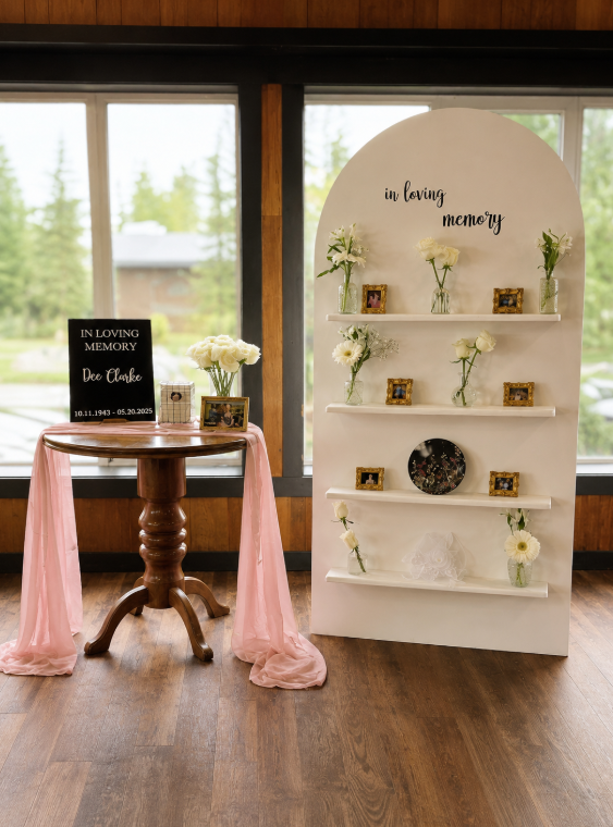 Memorial display with a white arch-shaped shelf decorated with white flowers and framed photos, and a round black plaque, accompanied by a wooden table covered with pink fabric, holding a bouquet of white flowers, a framed photo, and a black memorial sign with white text.