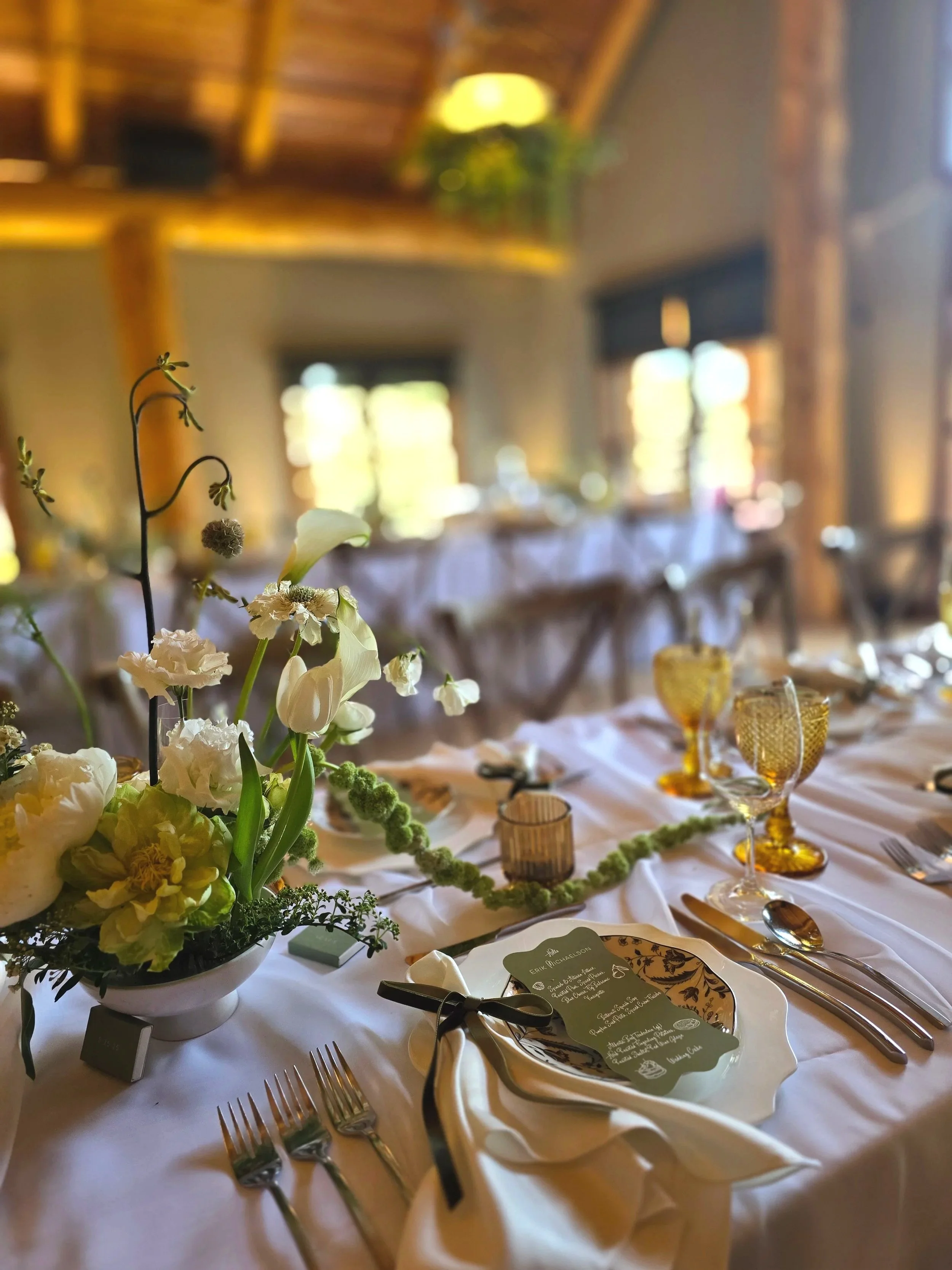 A close-up of a decorated banquet table with white tablecloth, floral centerpiece, glassware, and menu cards in a rustic-style venue with wooden beams and windows in the background.