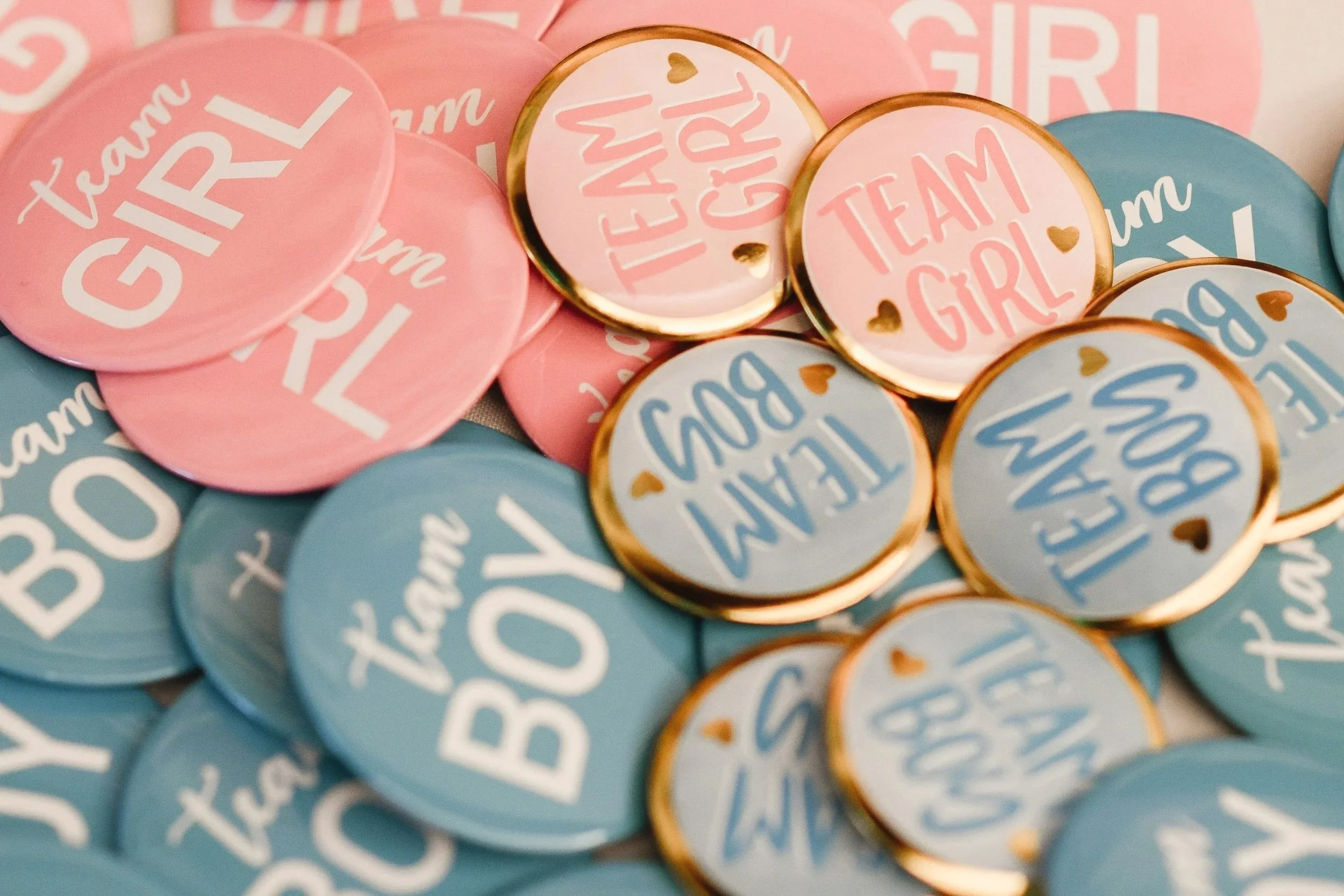 Close-up of round pink and blue pins with the words 'Team Girl' and 'Team Boy,' with small gold hearts.