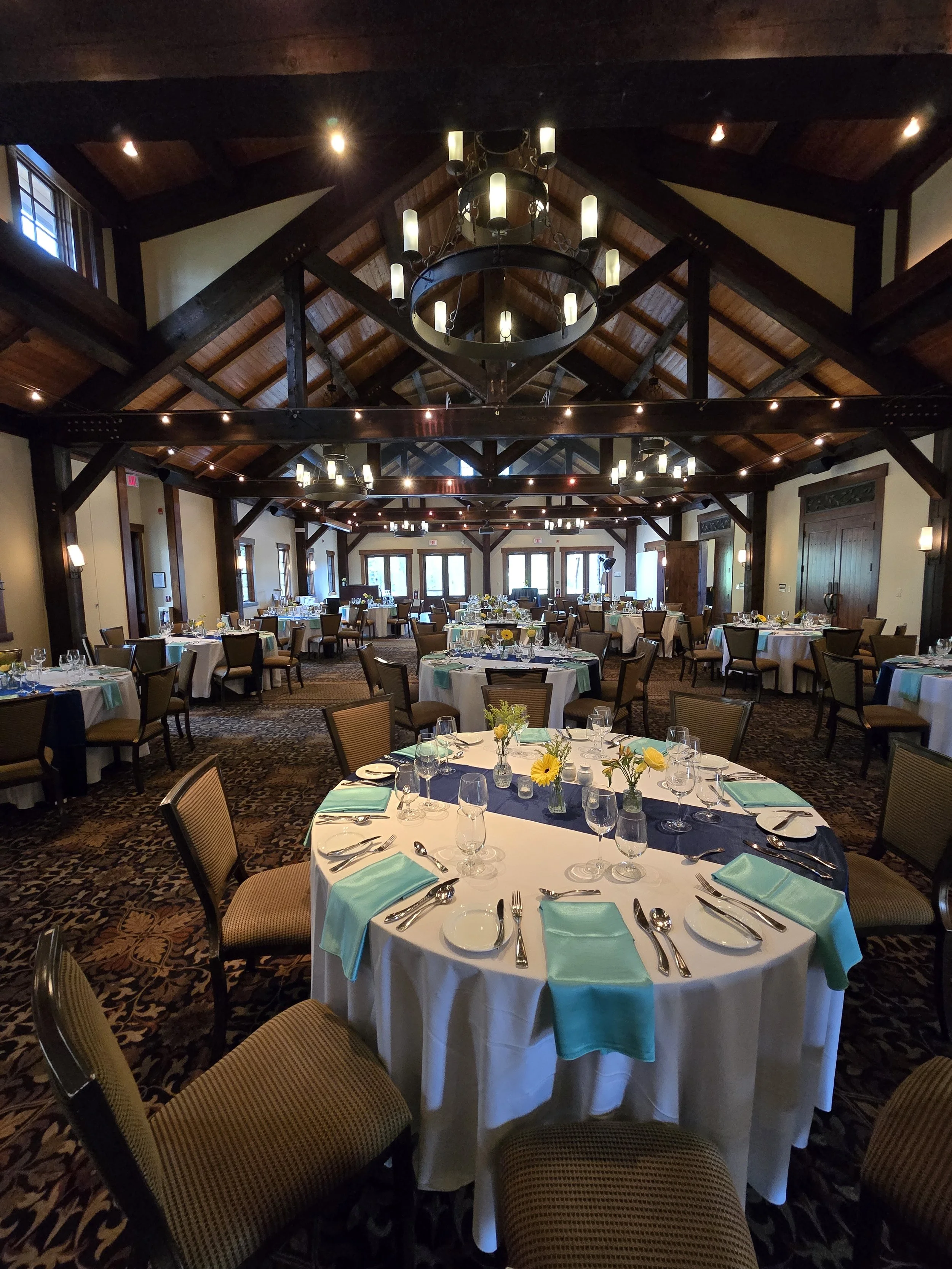 Set up banquet table with white tablecloths, turquoise napkins, glassware, silverware, and floral centerpieces in a decorated event hall with wood beams and chandeliers, ready for a wedding or formal celebration.
