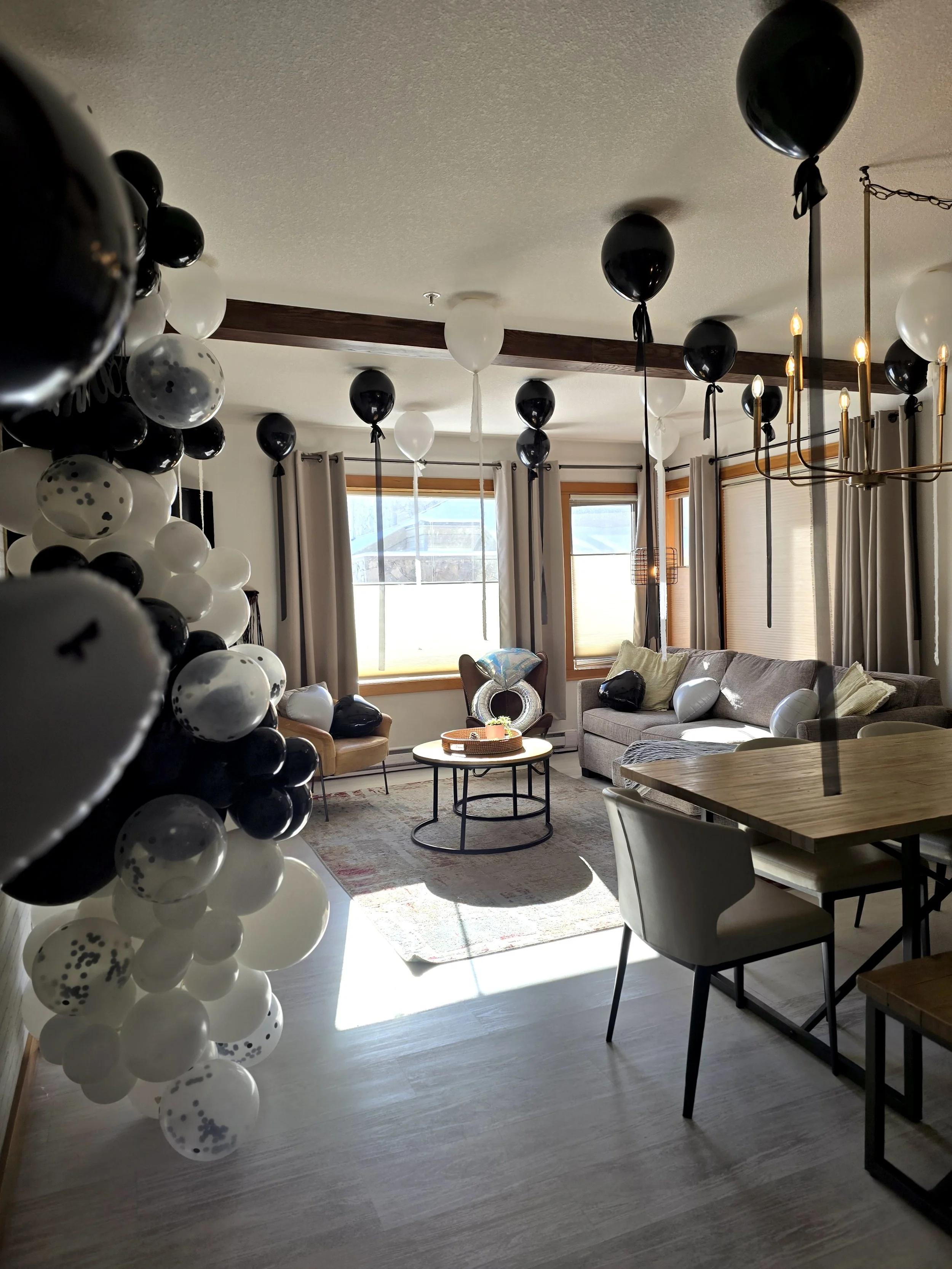 Living room decorated with black and white balloons, some with confetti, floating from the ceiling and scattered on furniture. There is a gray couch with pillows, a round coffee table, and a light-colored area rug. Sunlight streams through windows with white curtains.