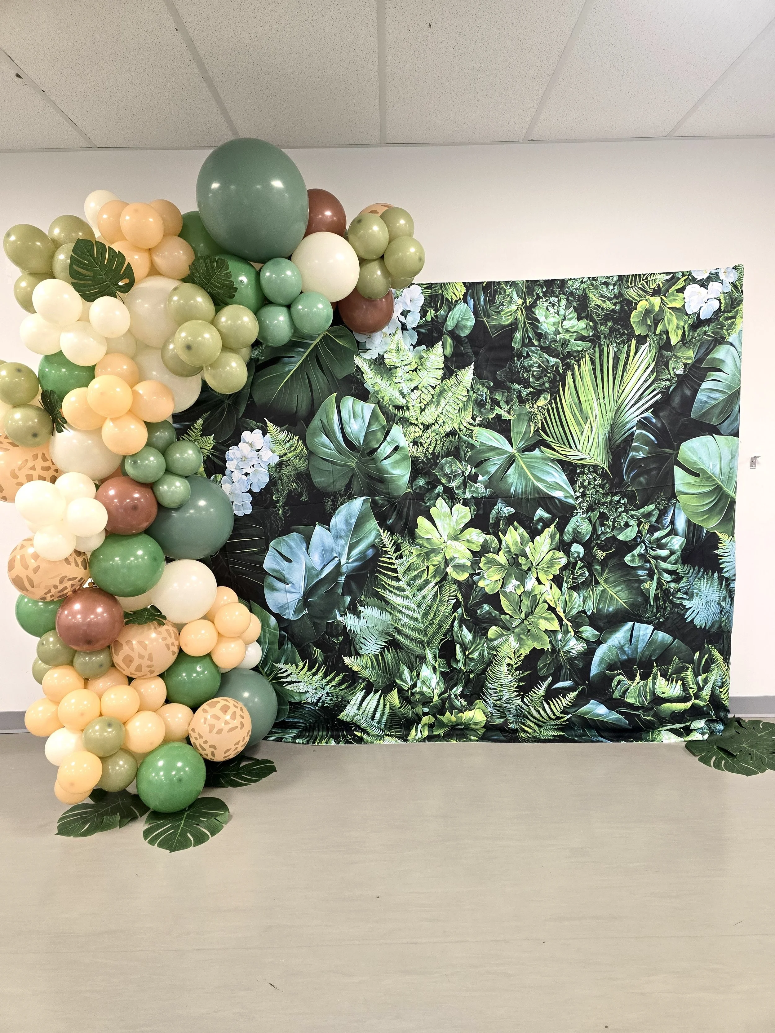 Decorative backdrop with green, beige, and brown balloons, and large green leaf accents, set against a leafy jungle-themed fabric background.