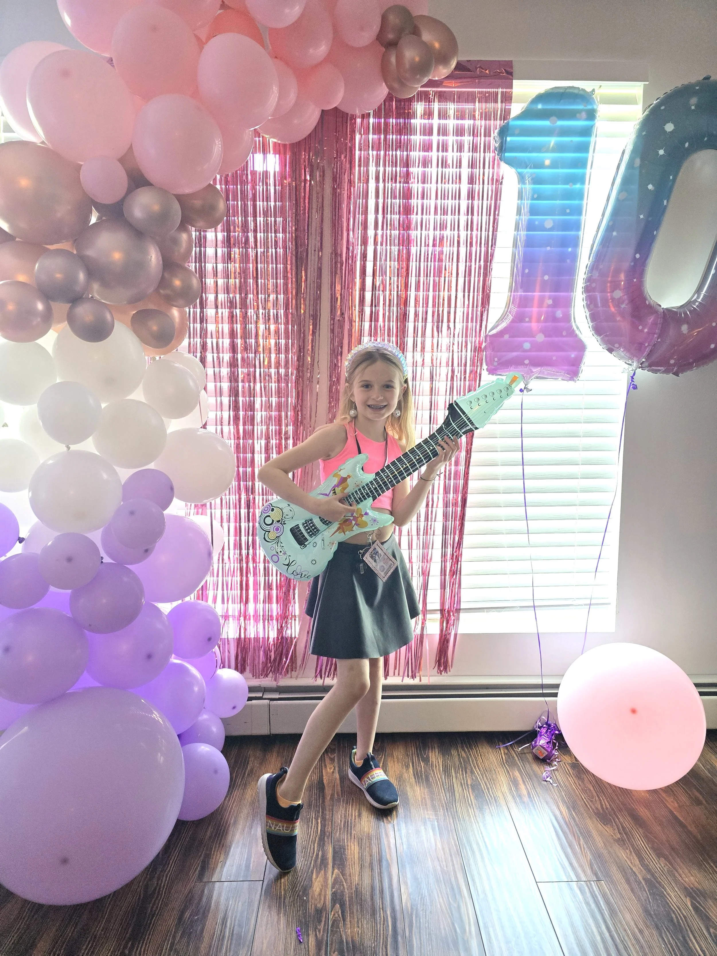 A young girl with blonde hair and a headband, wearing a pink top and a black skirt, holding a toy guitar, standing in front of pink and purple balloons, pink fringe curtain, and a large blue and pink balloon shaped as the number '1', with light coming through window blinds in the background.