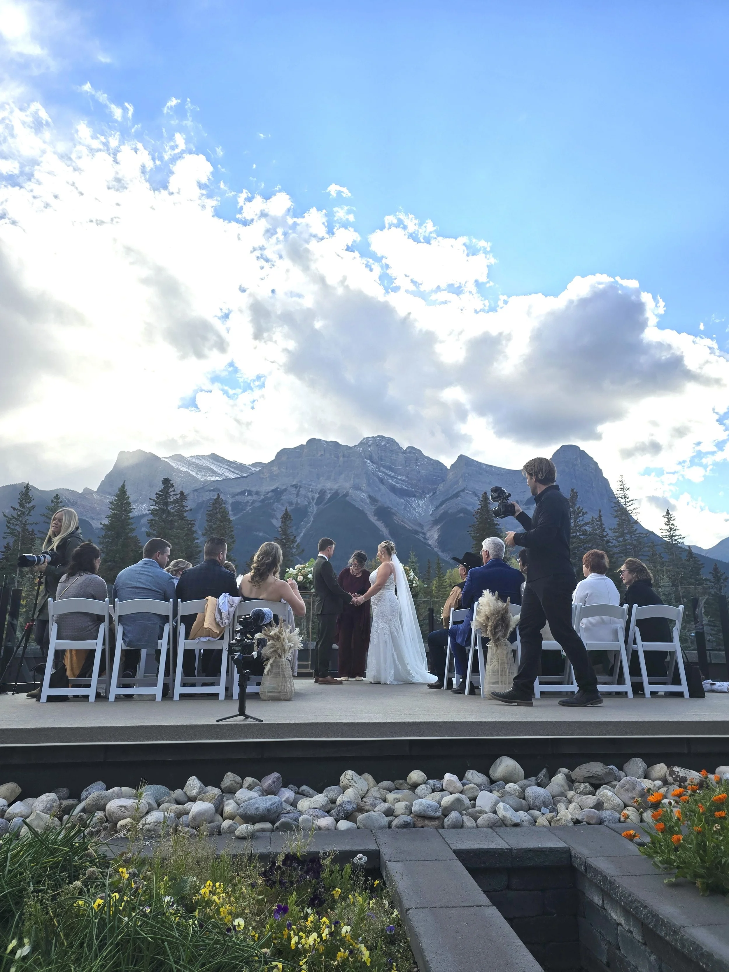 A wedding ceremony taking place outdoors in front of a mountain range under a blue sky with clouds. The bride and groom are standing in front of an officiant, surrounded by seated guests. A photographer is capturing the moment.