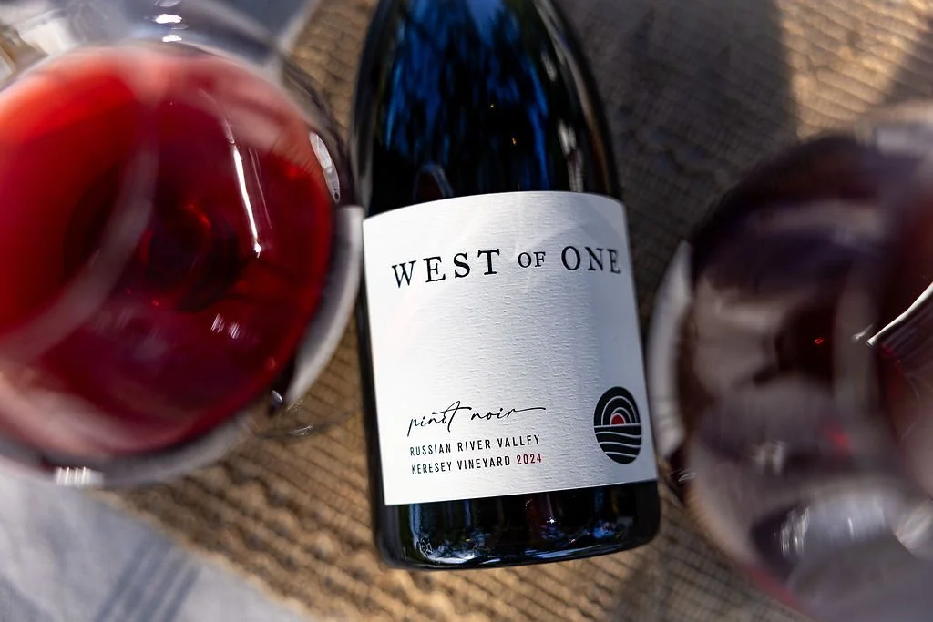 A bottle of West of One Pinot Noir wine from Russian River Valley Keresey Vineyard 2024, with two glasses of red wine on either side, on a textured surface.