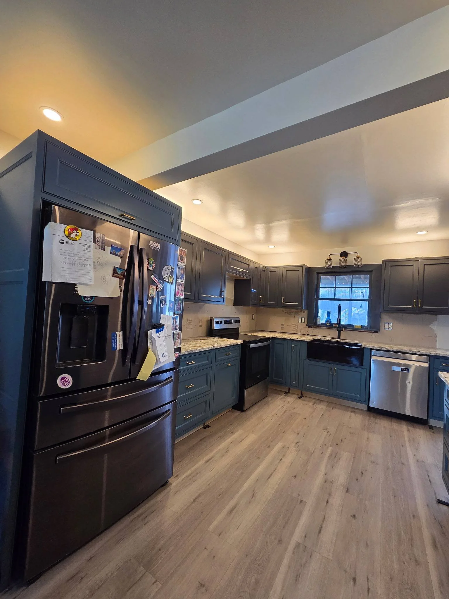 Modern kitchen with dark blue cabinets, stainless steel refrigerator, dishwasher, oven, and light wooden flooring. There is a window above the sink and ceiling lights.