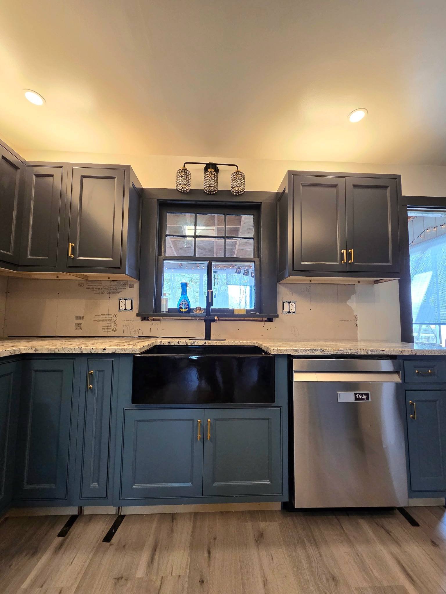 Modern kitchen with dark gray cabinetry, marble countertops, and a stainless steel dishwasher. There is a window above the sink, and the ceiling has recessed lighting and a decorative ceiling light fixture.