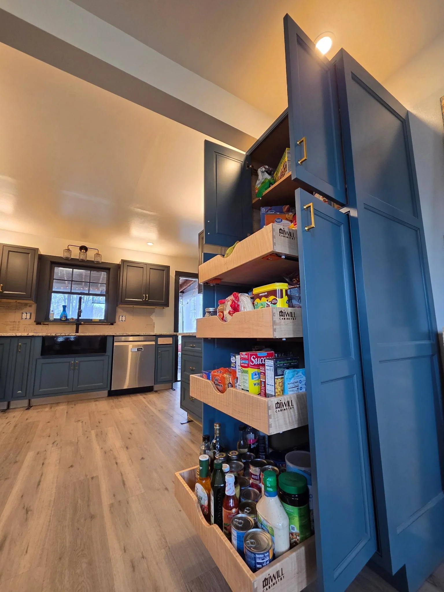 Open kitchen cabinet with snacks, drinks, and canned goods on pull-out shelves, with dark blue cabinetry and wooden flooring.