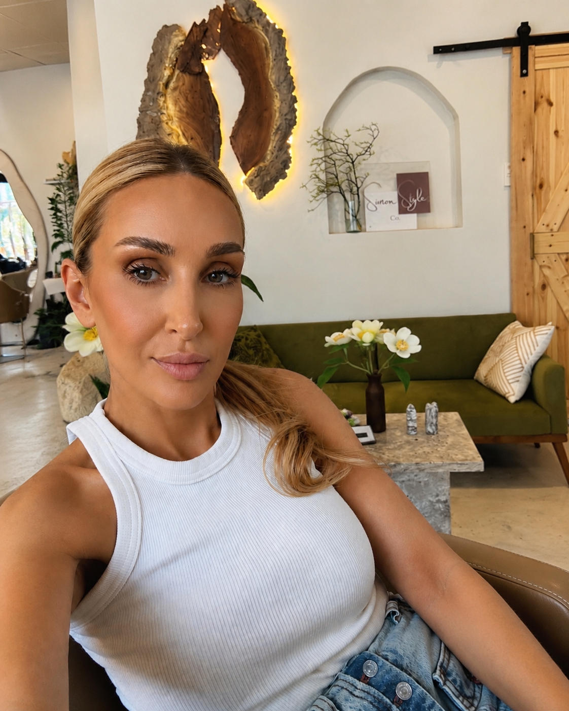 A woman with blonde hair and a white sleeveless top sitting on a chair in a stylish, cozy cafe with floral decor, a green sofa, and wooden accents.