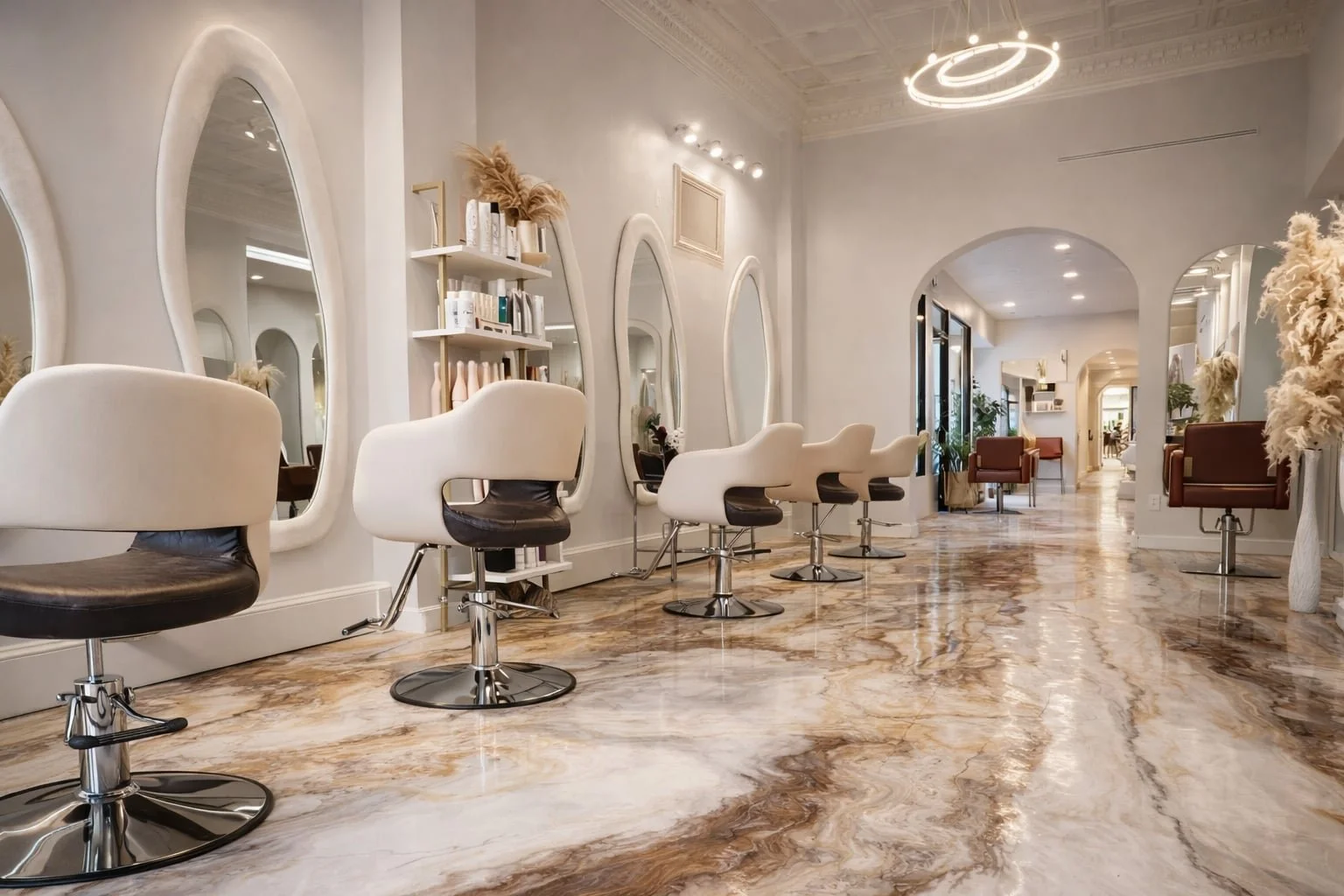 A modern hair salon with beige and brown salon chairs, large oval mirrors, floral decor, and a glossy marble floor.