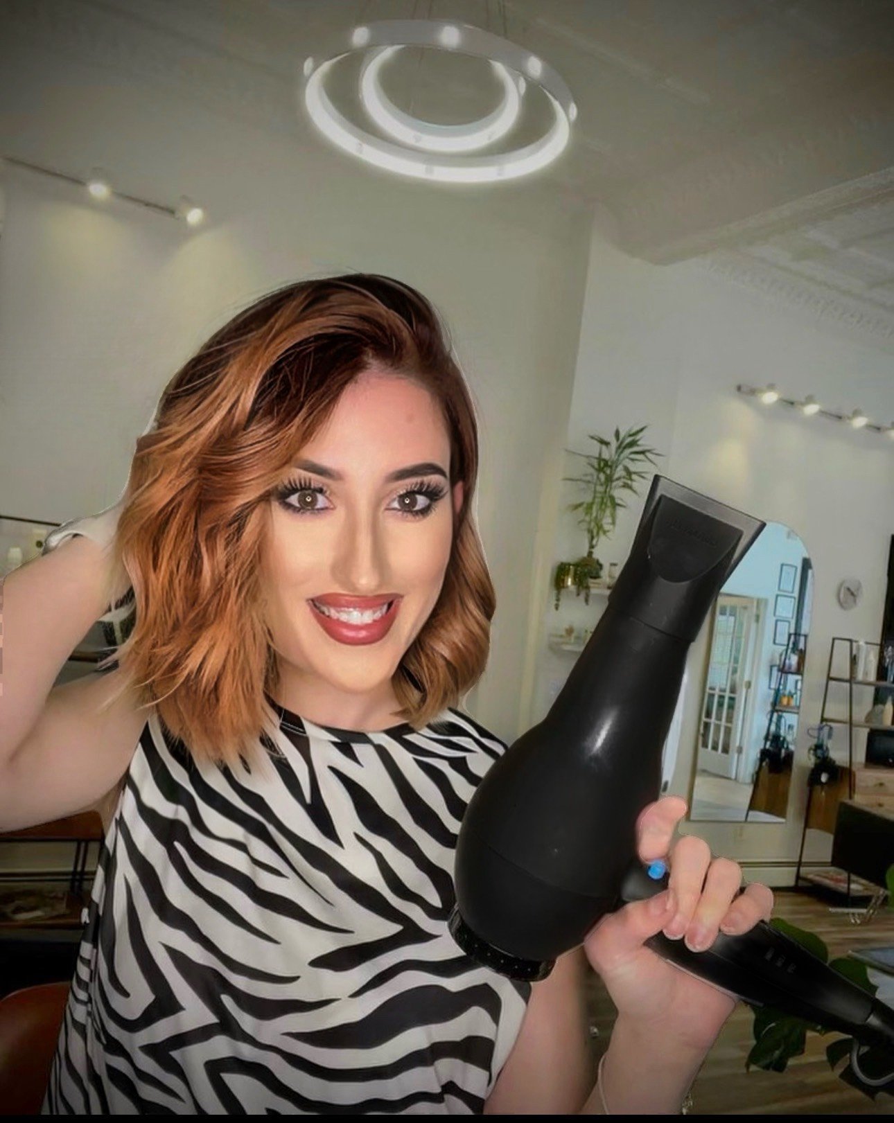 Woman in a salon holding a hairdryer, wearing a zebra-patterned top, with styled shoulder-length hair and makeup.