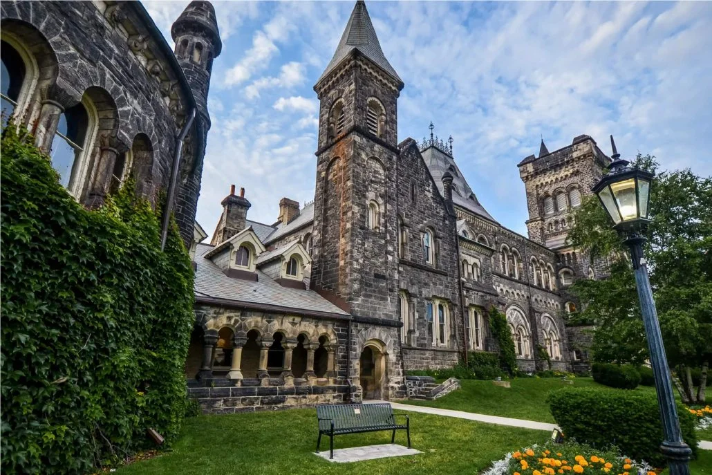 A romatic era building in brown-grey cobblestone with a green lawn and ivy growing up the fence. A lamppost adorns the front right of the photo.