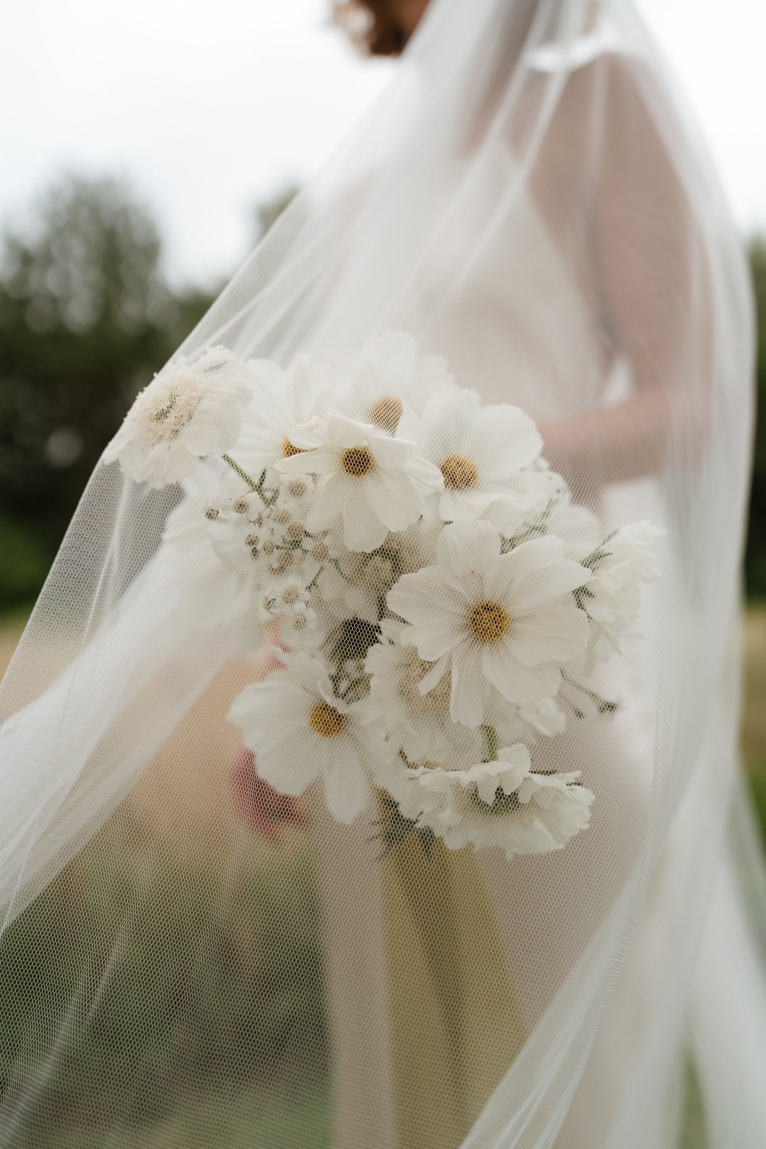 Close-up of a bride holding a bouquet of white flowers, partially covered by a sheer veil, outdoors.