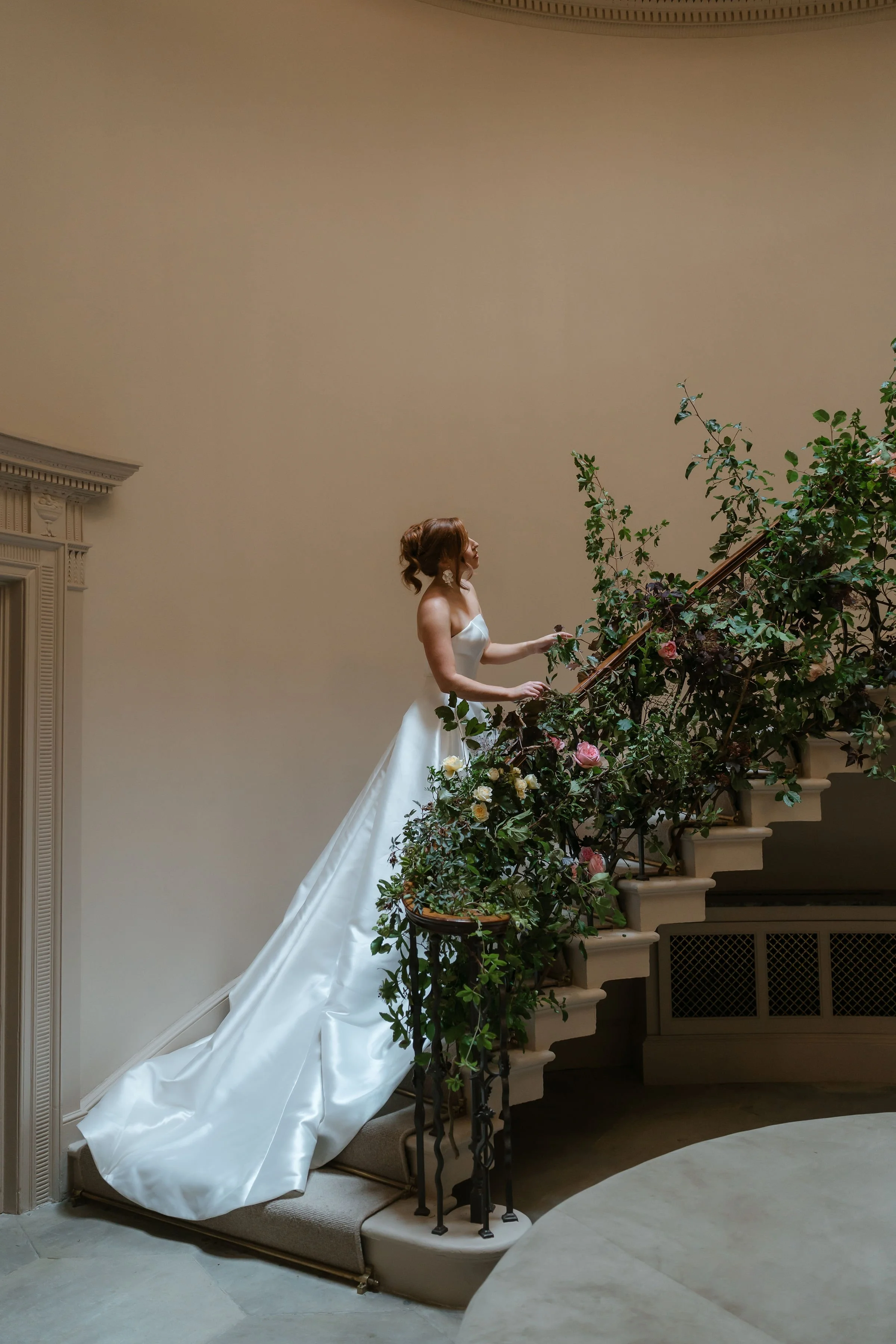 A woman in a white wedding dress stands on the staircase of Denton Reserve decorated with greenery and flowers.