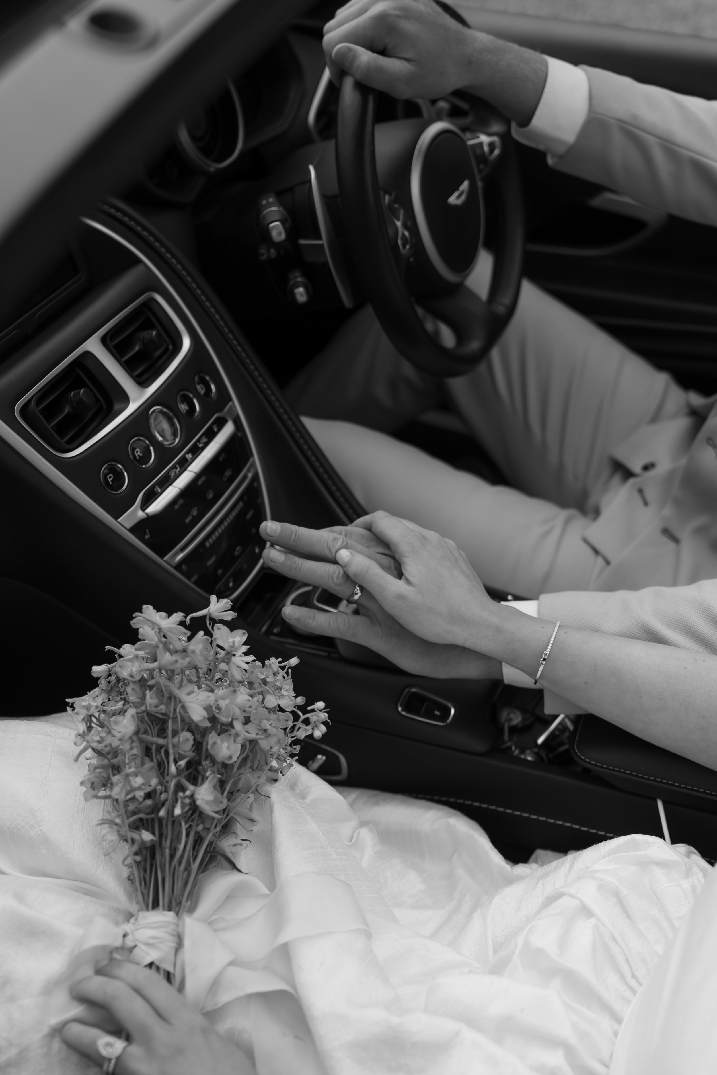 A black and white photo of a newlywed couple inside a car. The bride is holding a bouquet of flowers in her lap, and her hand is resting on the groom's hand which is on the car's gear shift. The groom has his hand on the steering wheel, both are dres
