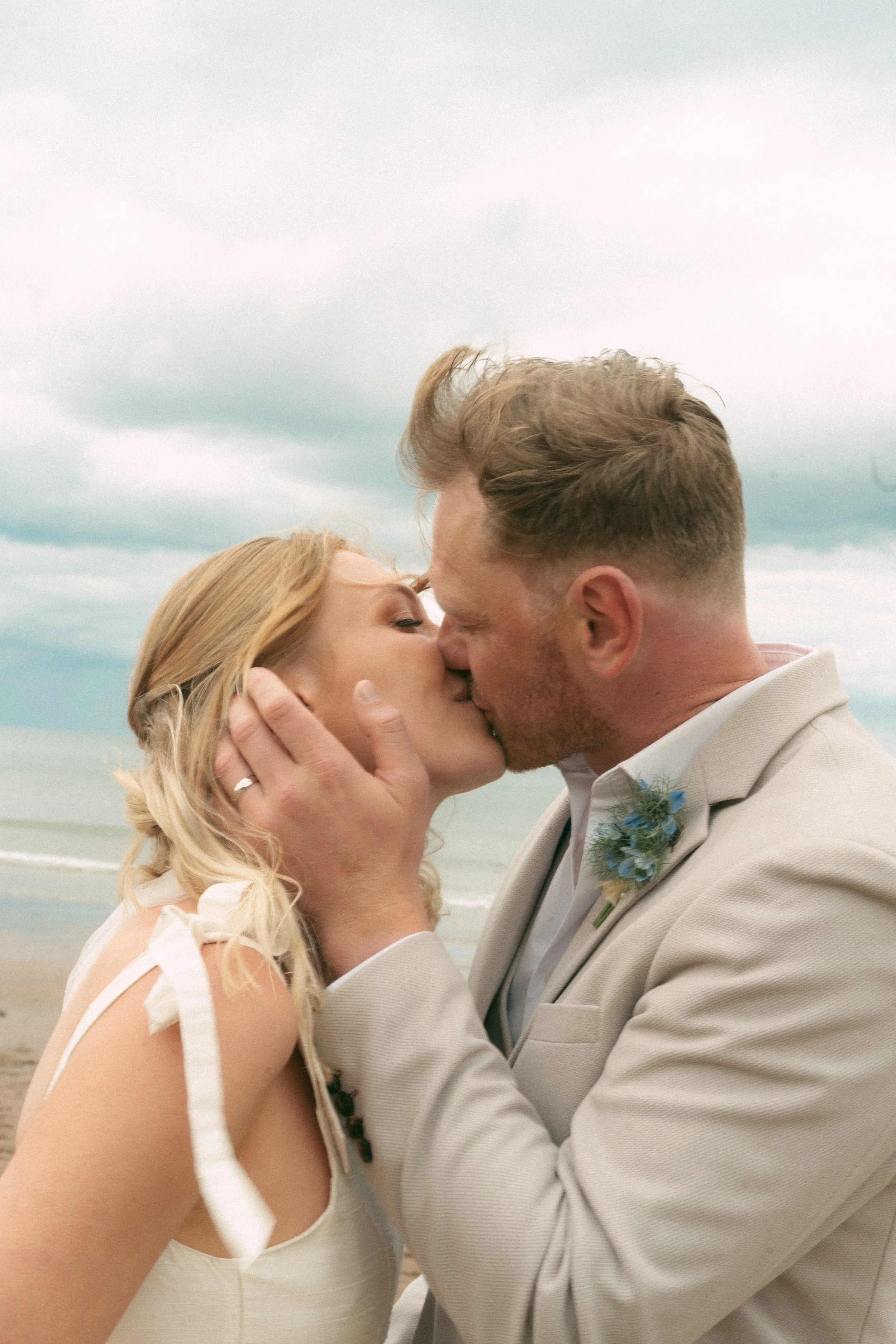 A couple kissing on the beach, the woman with blonde hair and the man with short brown hair, wearing wedding attire, with cloudy sky in the background.