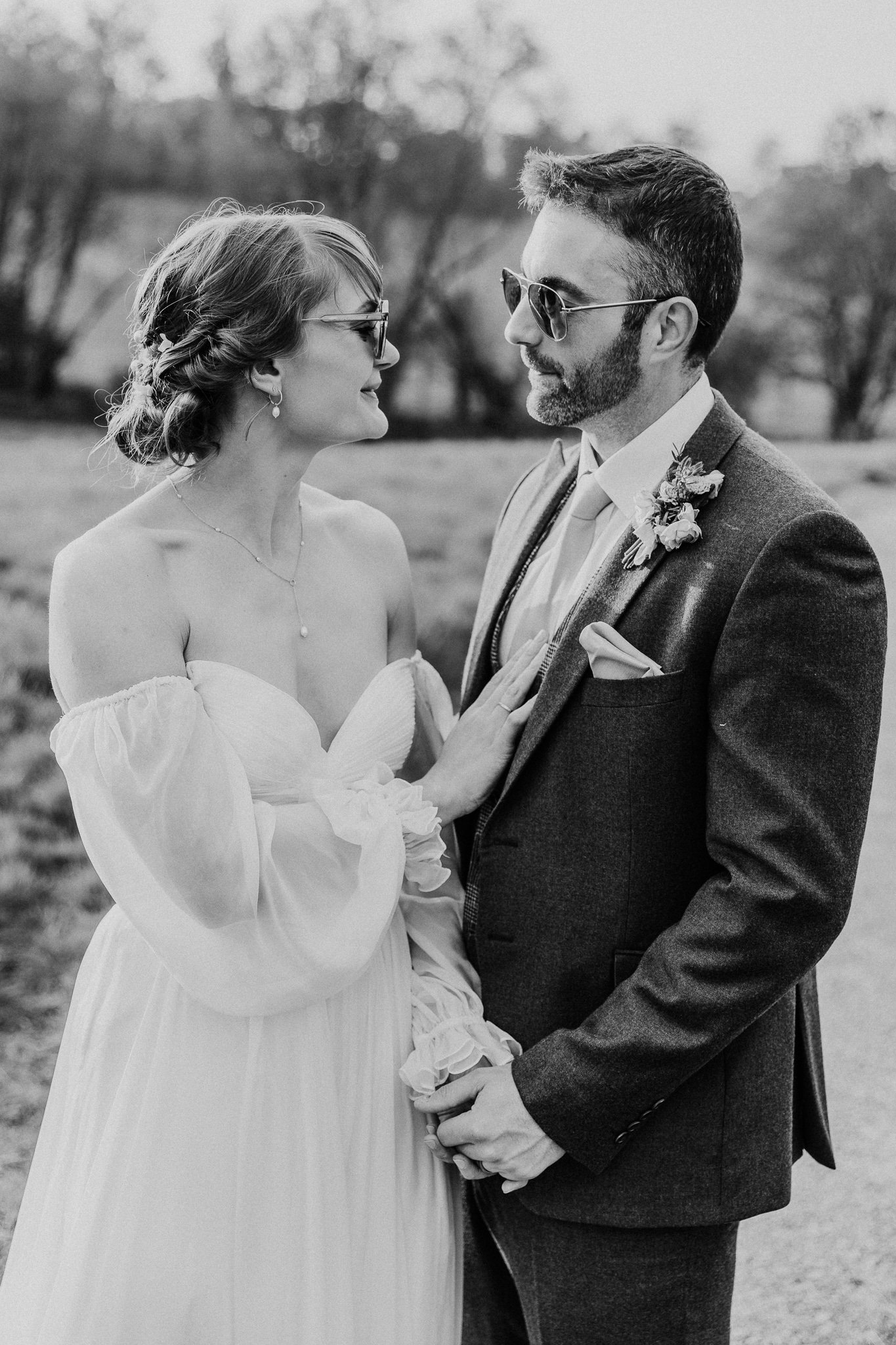Black and white photo of a bride and groom standing close together outdoors. The bride is wearing a strapless wedding dress and glasses, and the groom is in a suit with sunglasses. They are looking at each other lovingly.
