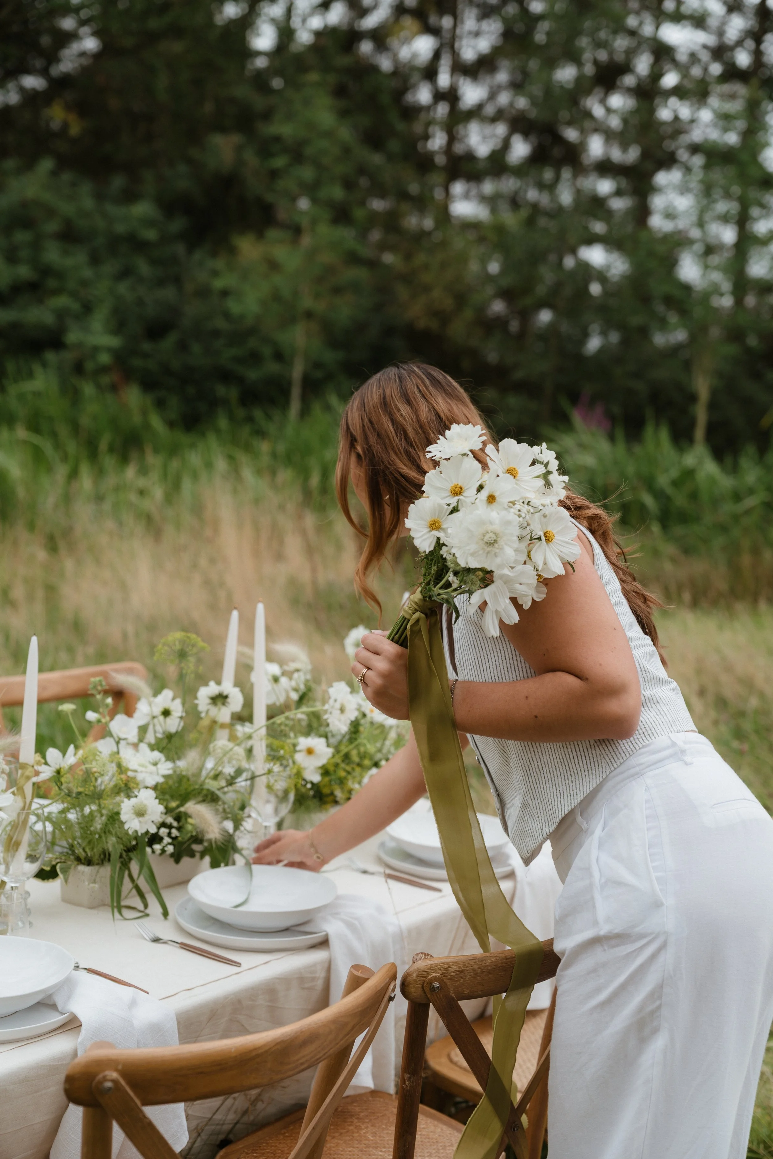A woman with long wavy hair, holding a bouquet of white flowers and leaning over a decorated table outdoors in a grassy area with trees in the background.