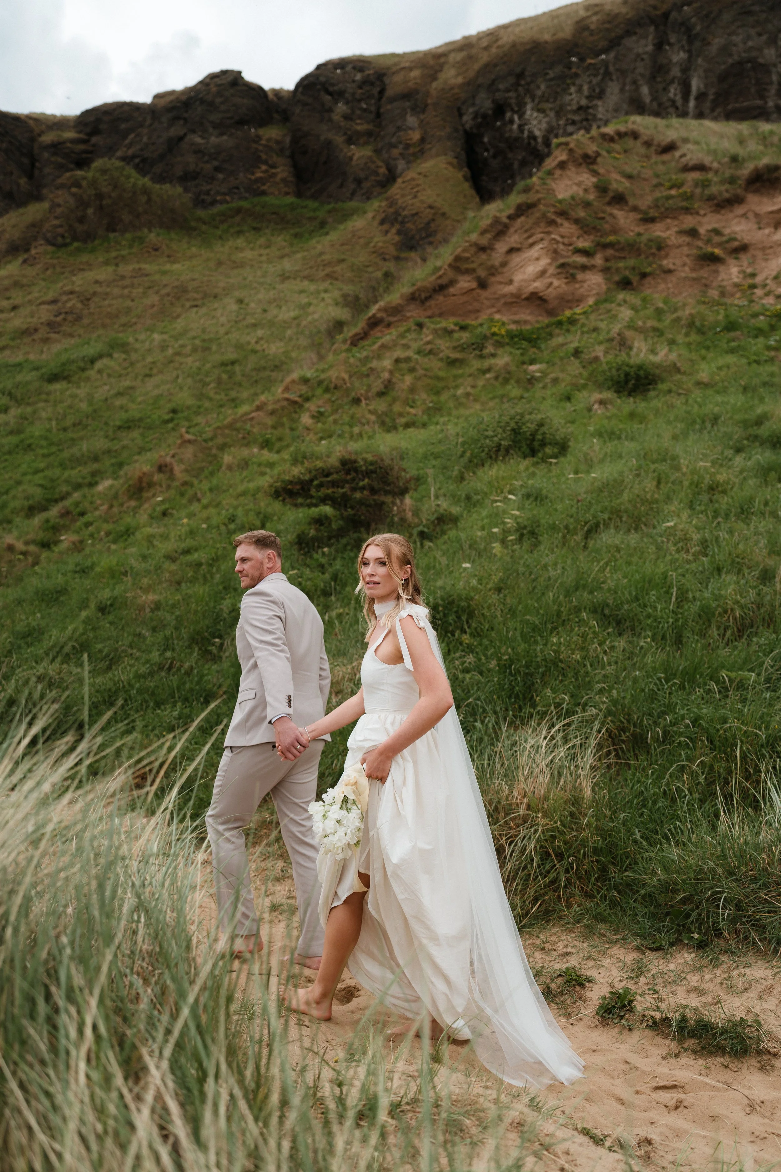 A couple walk through the sand dunes on their wedding day on a beach in Yorkshire. The bride wears a wedding dress that she lifts at the knee whilst holding a bouquet of sweetpeas.