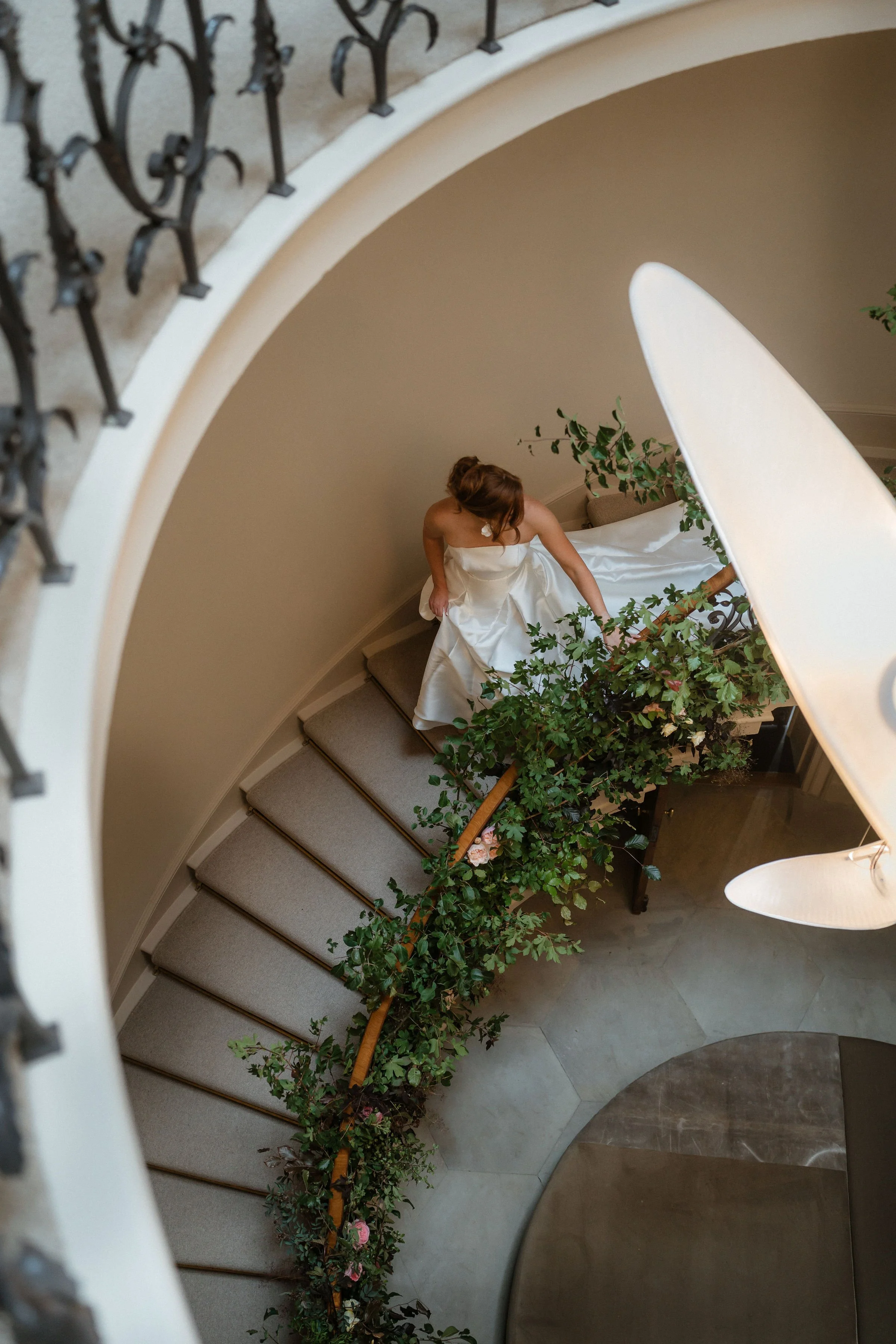 A woman in a white gown standing on the staircase of Denton Reserve, reaching for a plant on the railing, viewed from above through a spiral staircase balustrade.